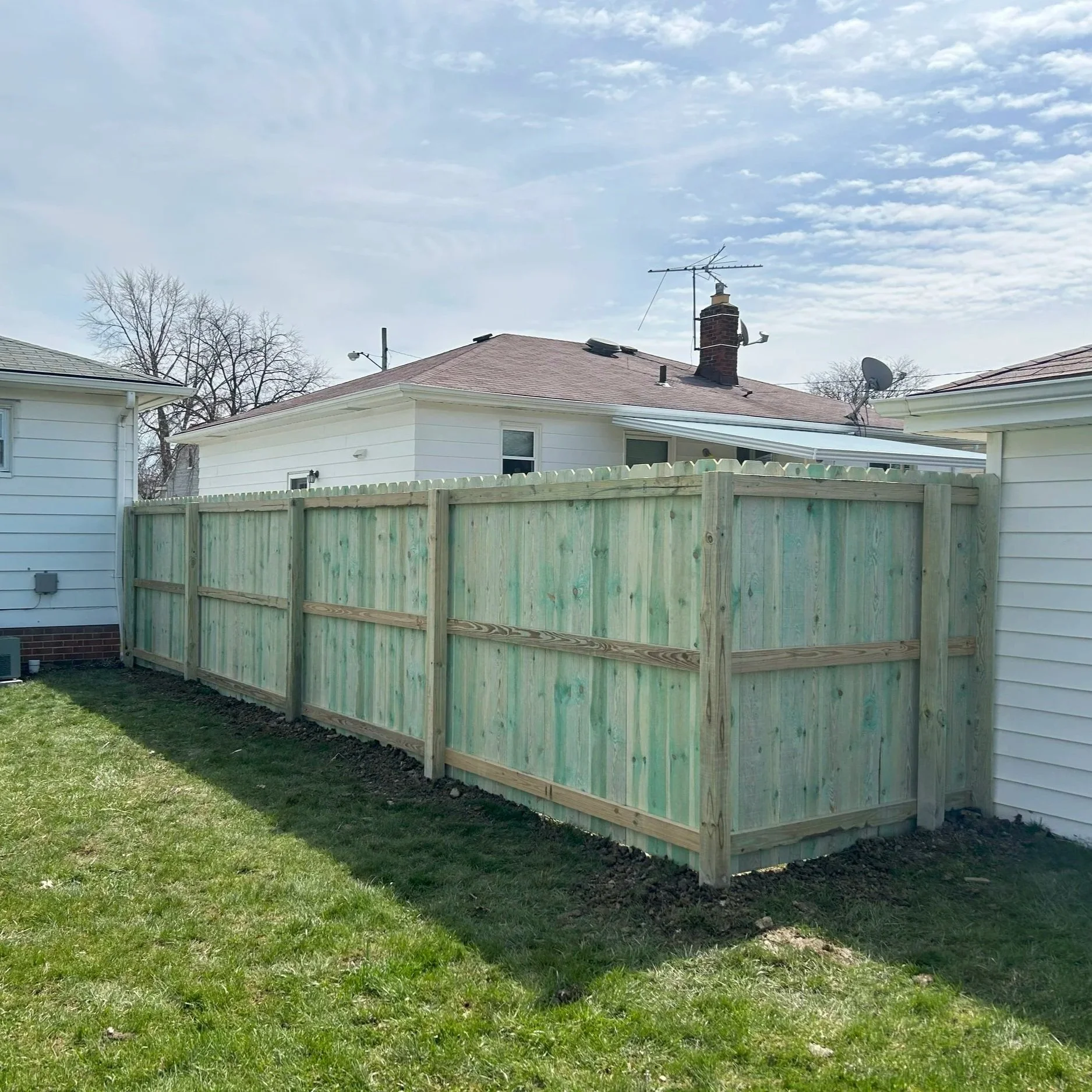 Newly built wooden fence in a backyard, with grass in the foreground and neighboring houses in the background under a partly cloudy sky.