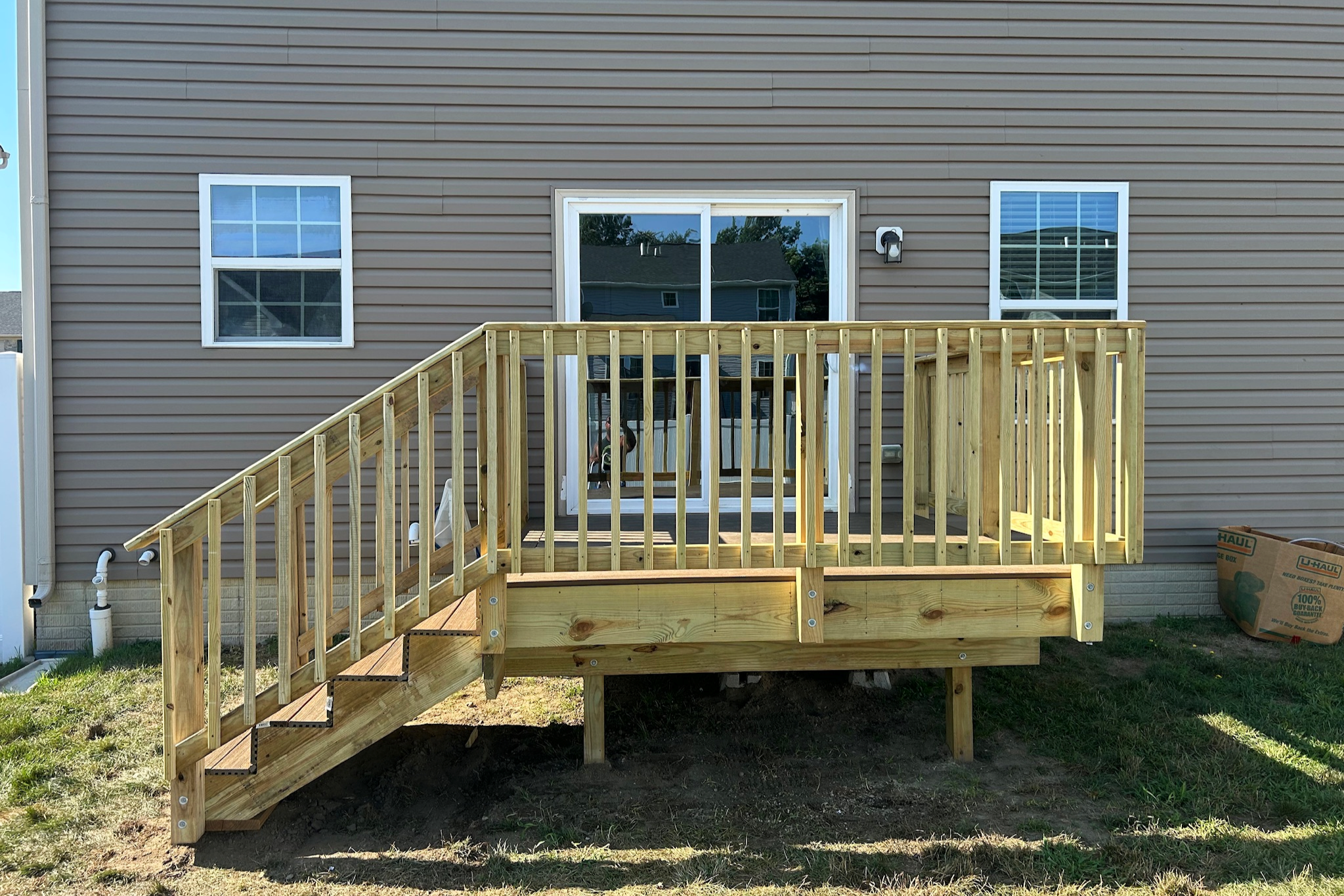 New wooden deck with stairs and railing attached to the back of a beige house with three windows and sliding glass door.