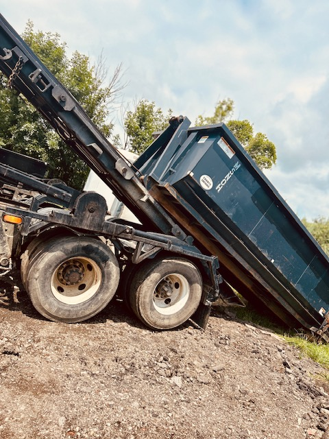 A dump truck with its bed raised, spilling asphalt or gravel onto the ground, on a construction site with trees in the background.