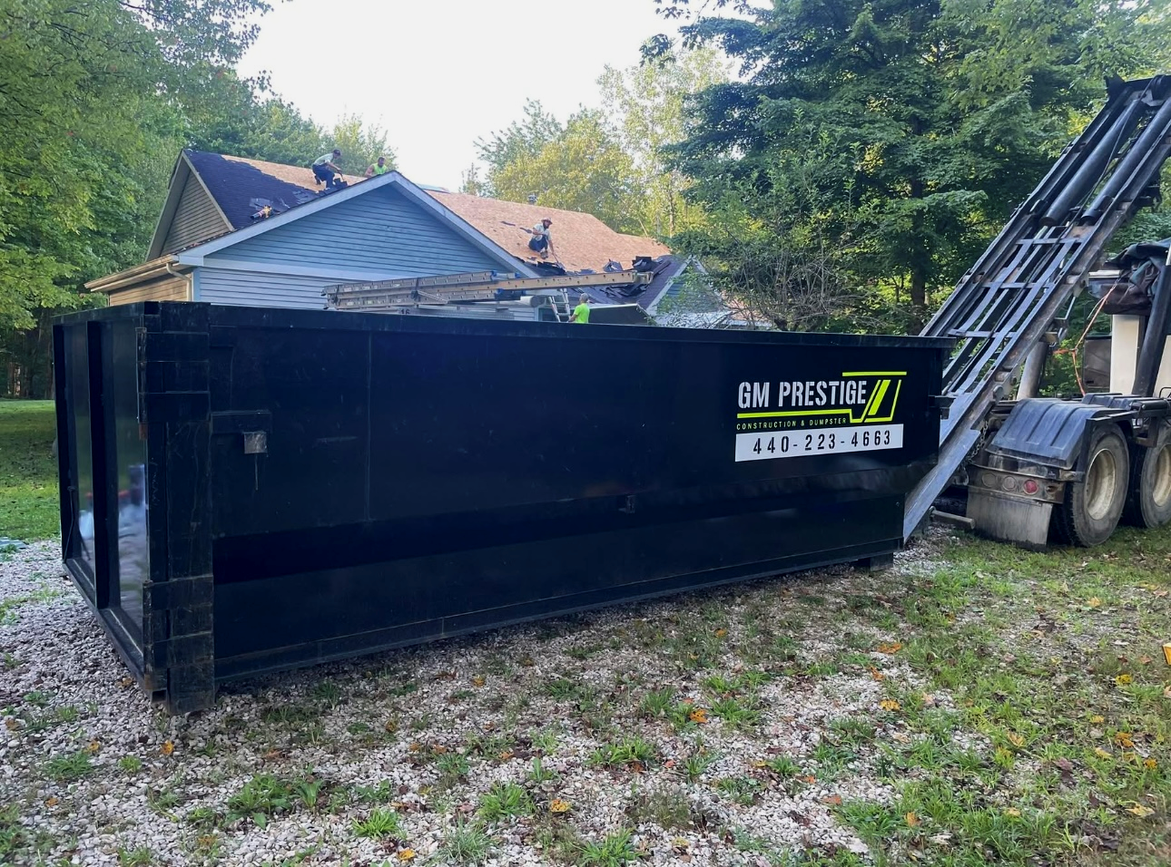 Construction workers working on roof of a house with a blue exterior, using a crane and a large black dumpster nearby labeled 'GM Prestige' for debris.