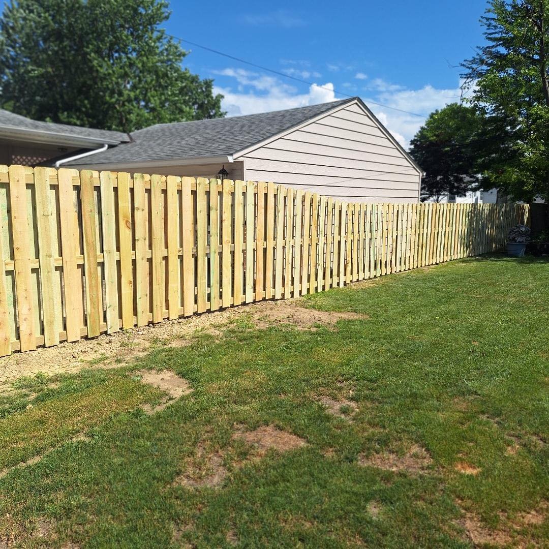 A newly built wooden privacy fence surrounds a backyard with green grass, a house with beige siding, and a blue sky with some clouds.
