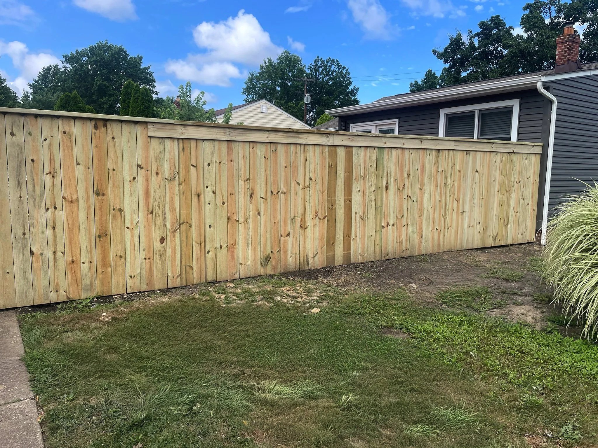 A newly installed wooden fence with vertical planks in a backyard next to a house with dark gray siding and a white-trim window, under a partly cloudy blue sky.