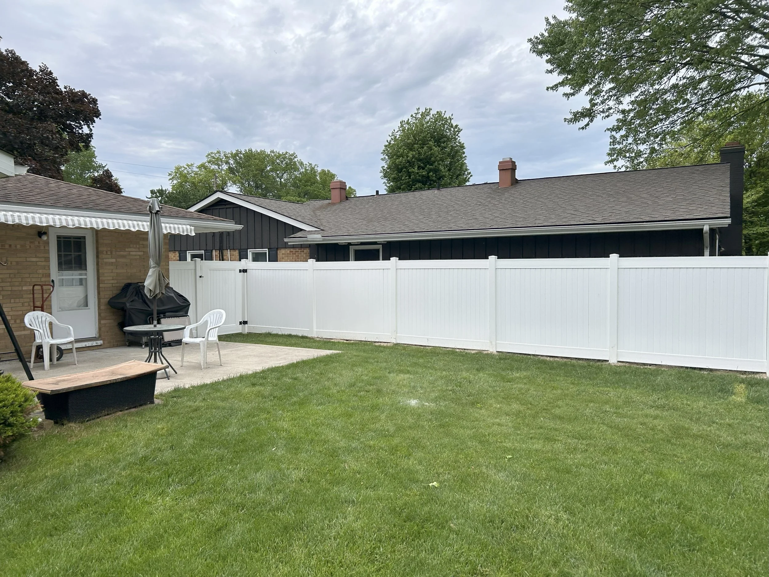 A backyard with a green lawn, a concrete patio, a table with an umbrella, and white plastic chairs. There is a white privacy fence and a brick house on the left side, with another black house in the background. Overcast sky.