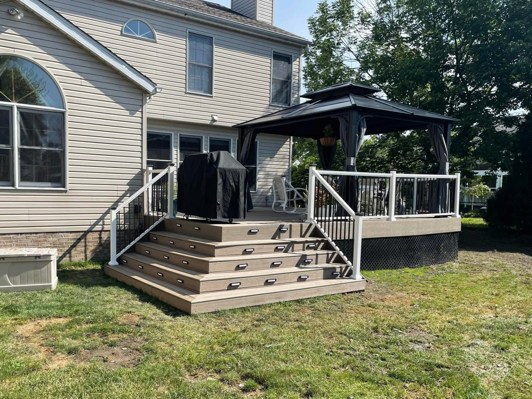 Backyard wooden deck attached to a house, with stairs leading up to a gazebo and a grill cover on the deck.