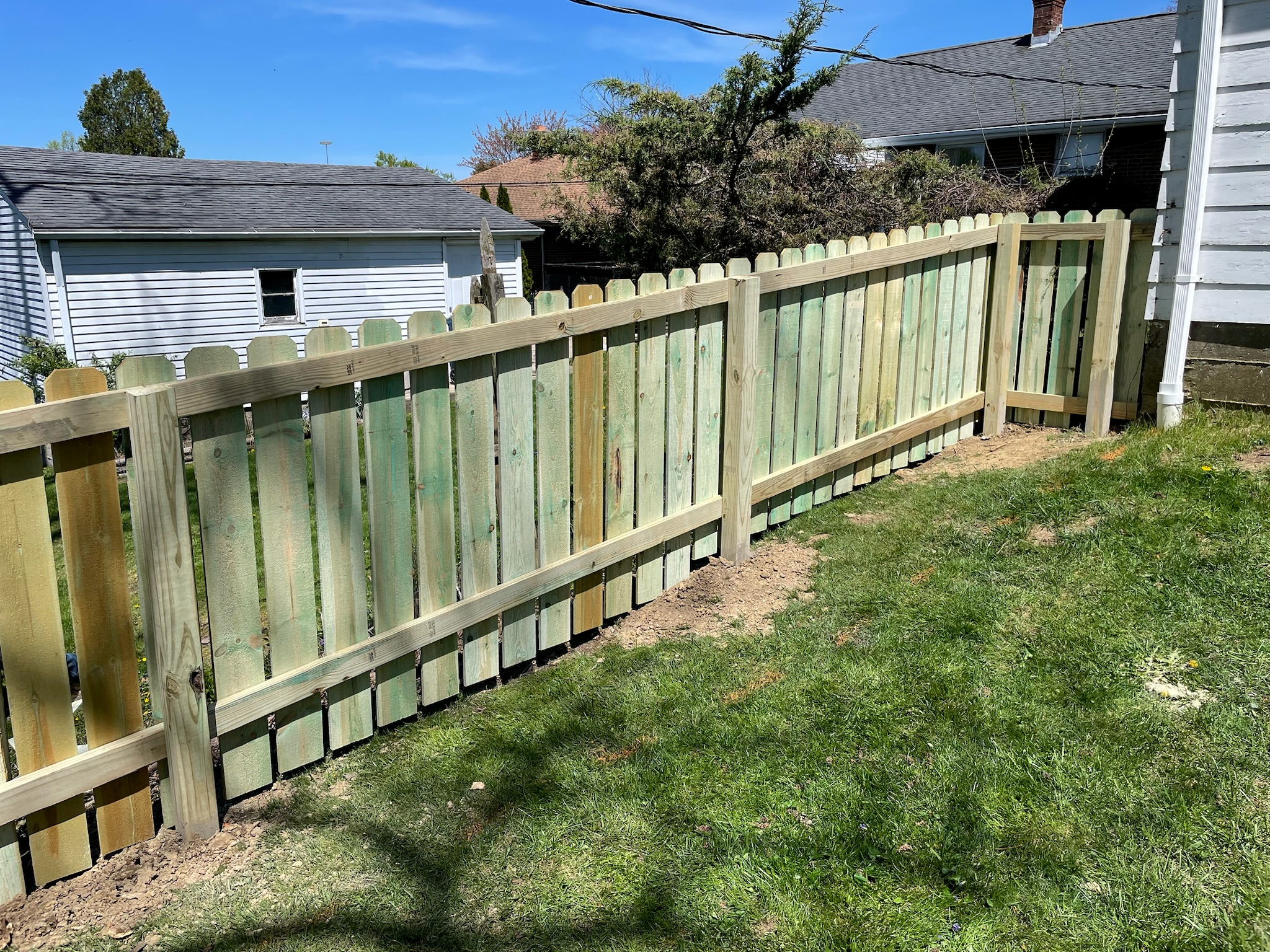 Newly built wooden fence in a backyard, with grass and dirt on the ground, and neighboring houses in the background under a clear blue sky.