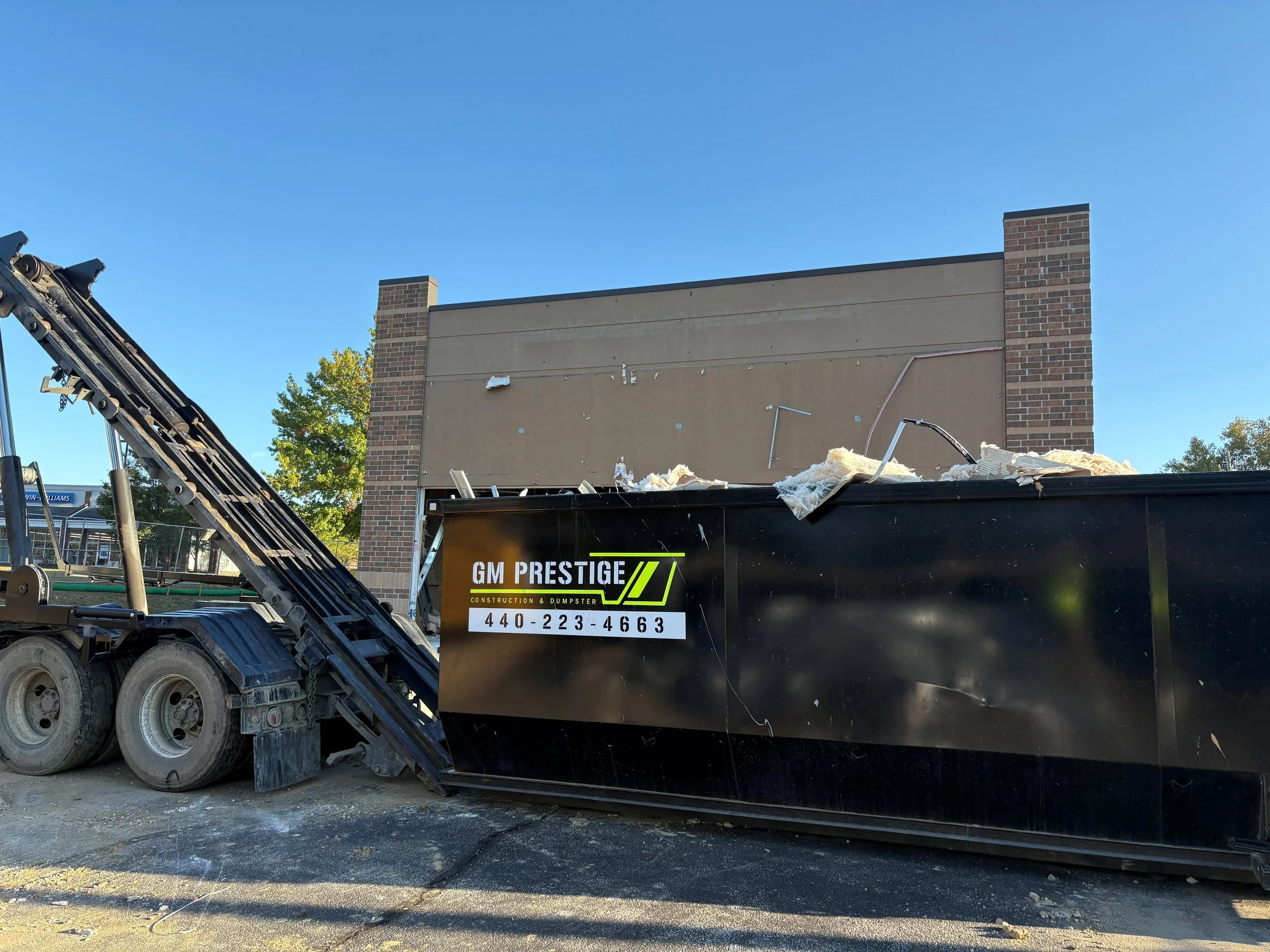 A construction dumpster overflowing with debris in front of a building undergoing renovation, with a black conveyor belt and construction truck nearby.