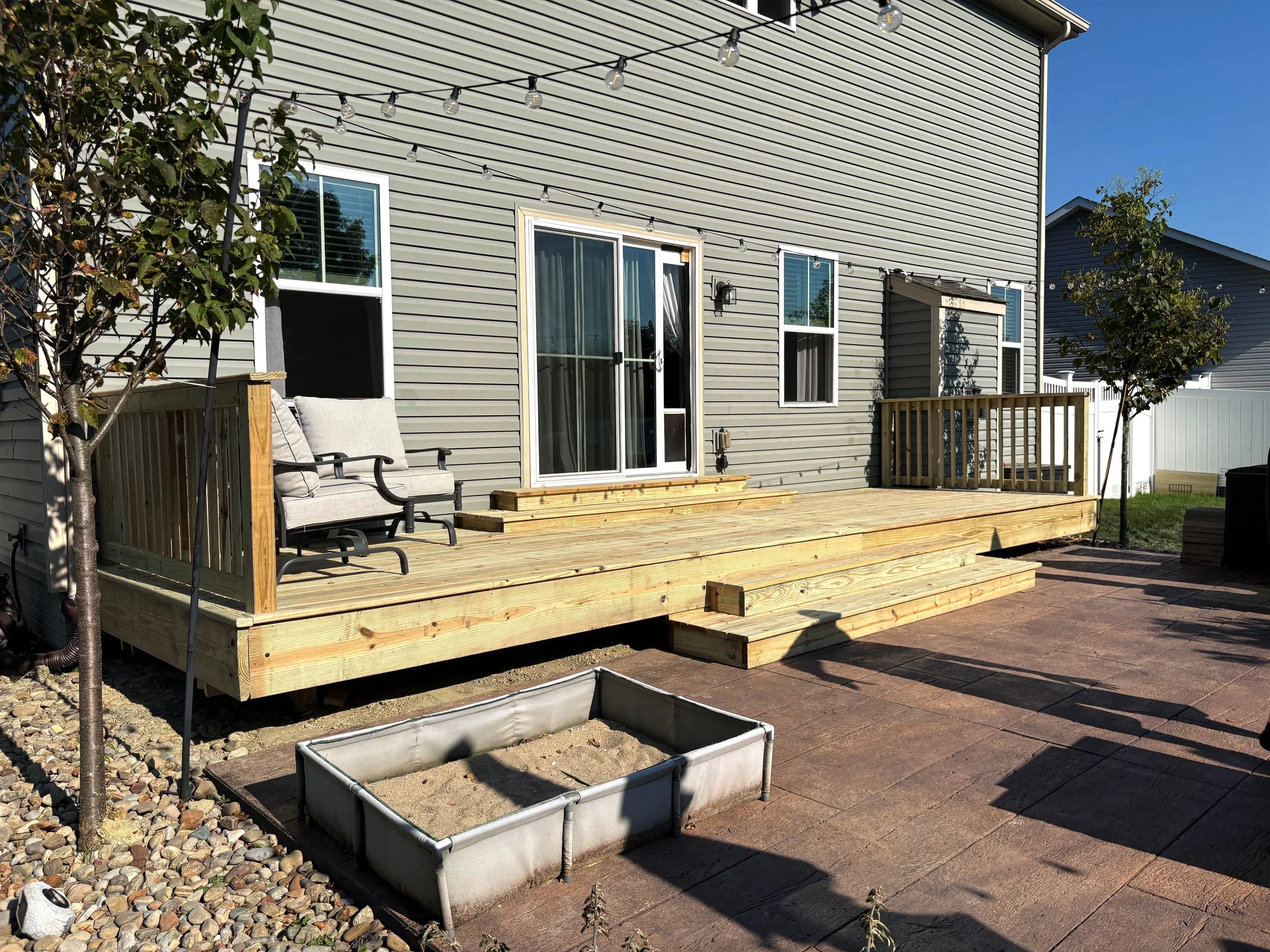 Backyard with a newly constructed wooden deck with stairs, two small trees, and a patio area with paving stones. There is outdoor seating on the deck, a pot with soil, and a white fence in the background.
