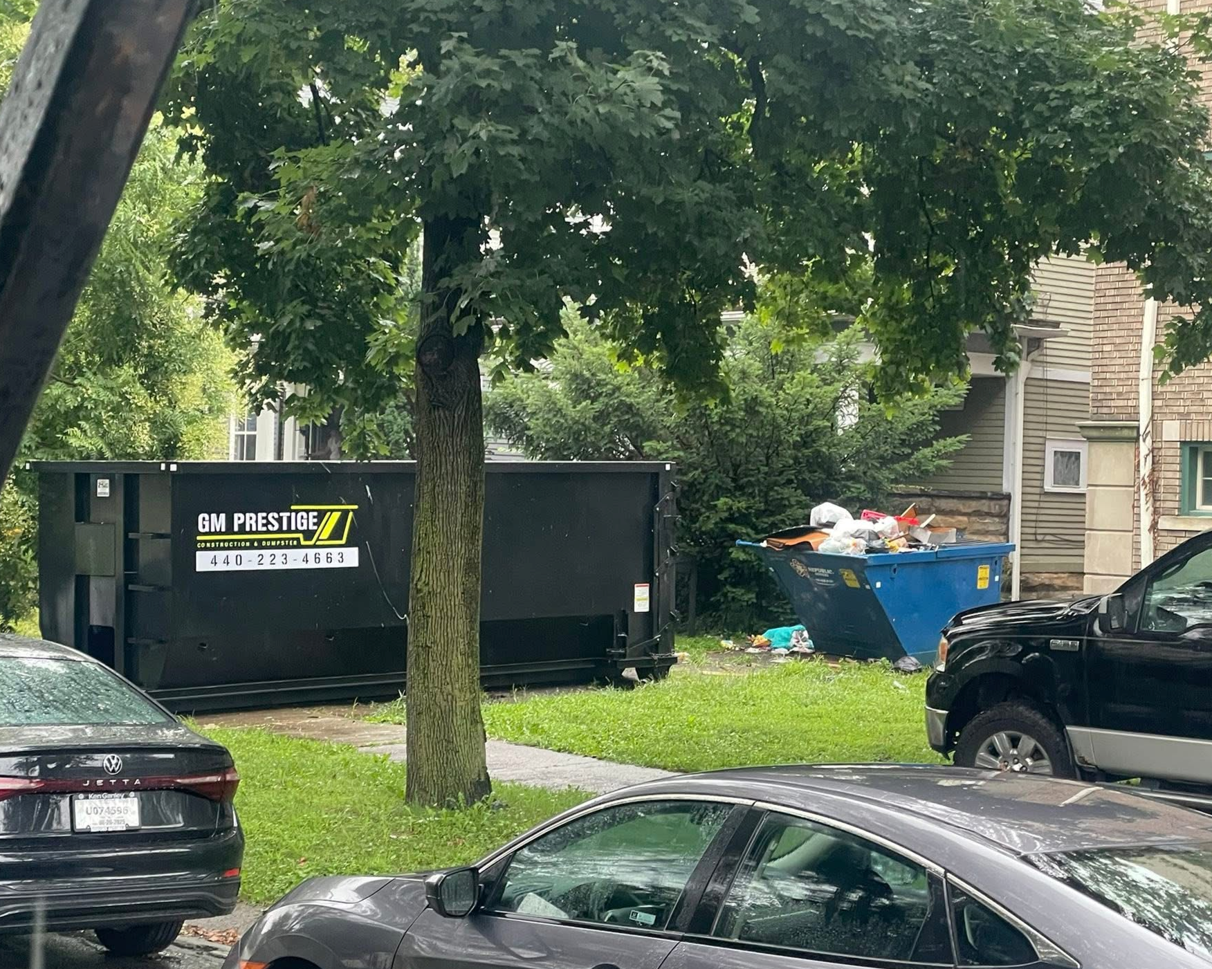 A residential street scene with cars parked along the curb, a green grassy lawn with a tree, a large black dumpster labeled GM Prestige, and a blue dumpster overflowing with trash next to a house with siding.