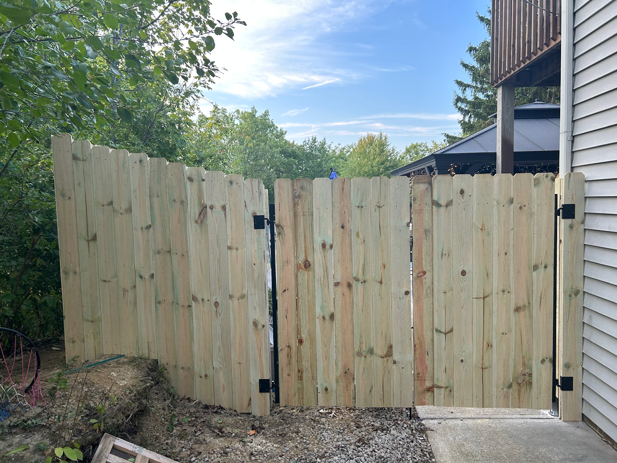 New wooden fence with gate next to house on right, greenery and trees in background, partly cloudy sky.