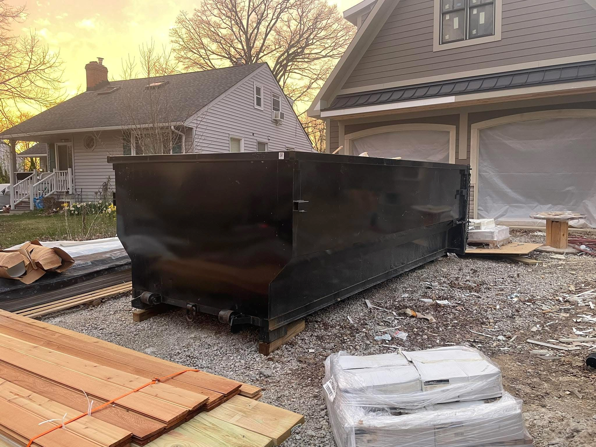 A construction site with building materials, including a large black metal dumpster, stacks of wood and wrapped insulation, in front of a house with construction wrapping on the windows during sunset.