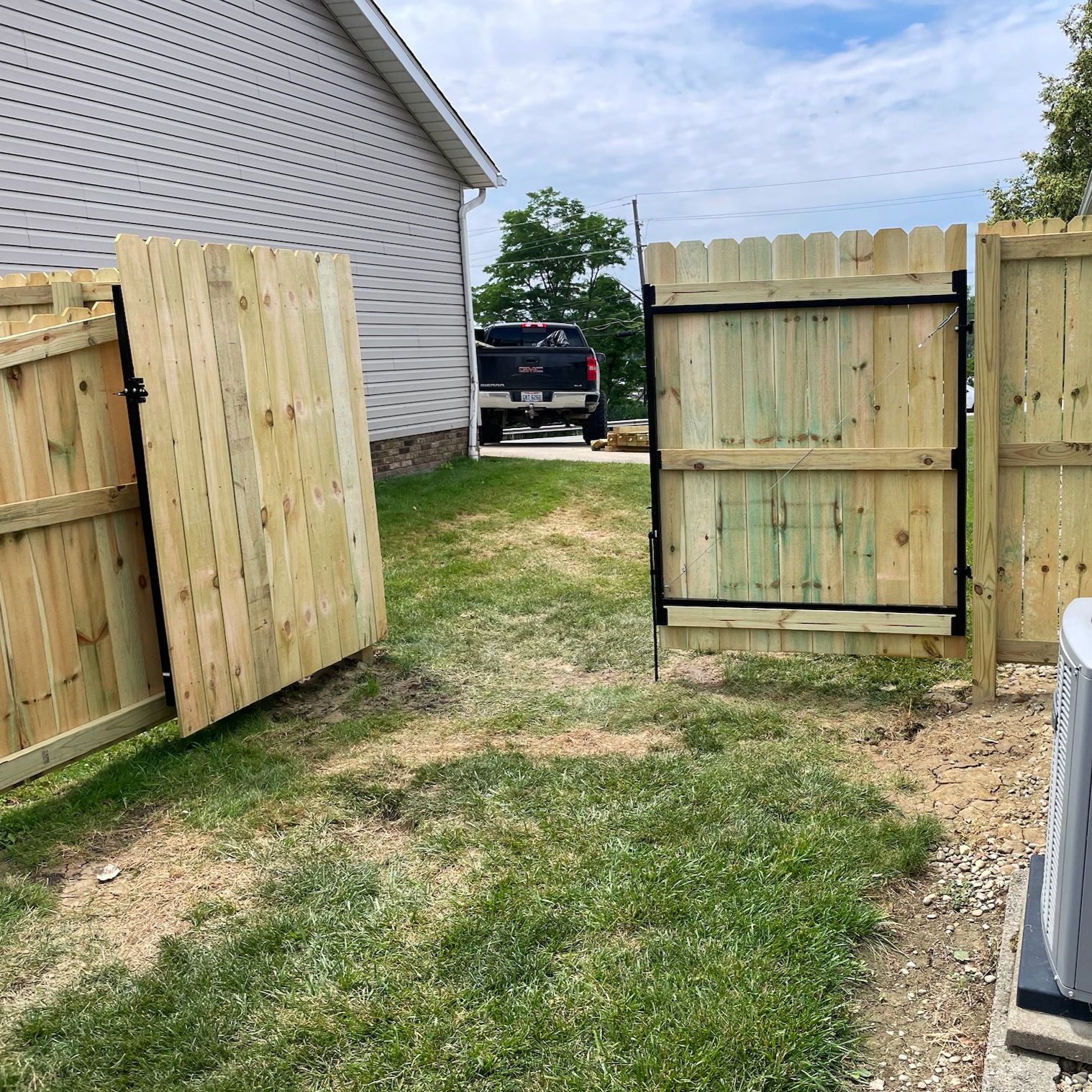 A partially open wooden gate with a latch, revealing a grassy backyard with a black pickup truck parked near a house with vinyl siding.