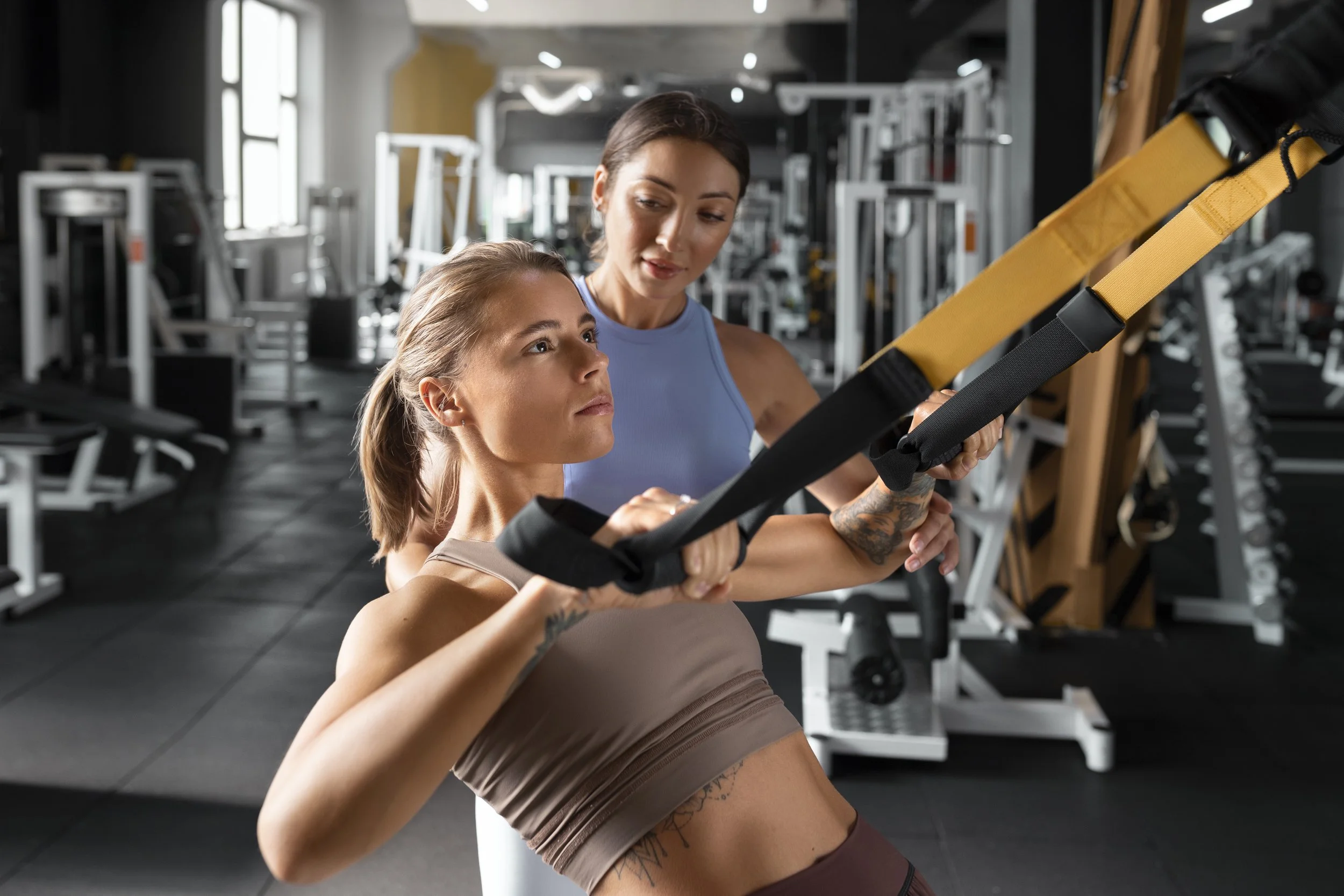 Two women in workout attire using a suspension training strap in a gym with exercise equipment in the background.