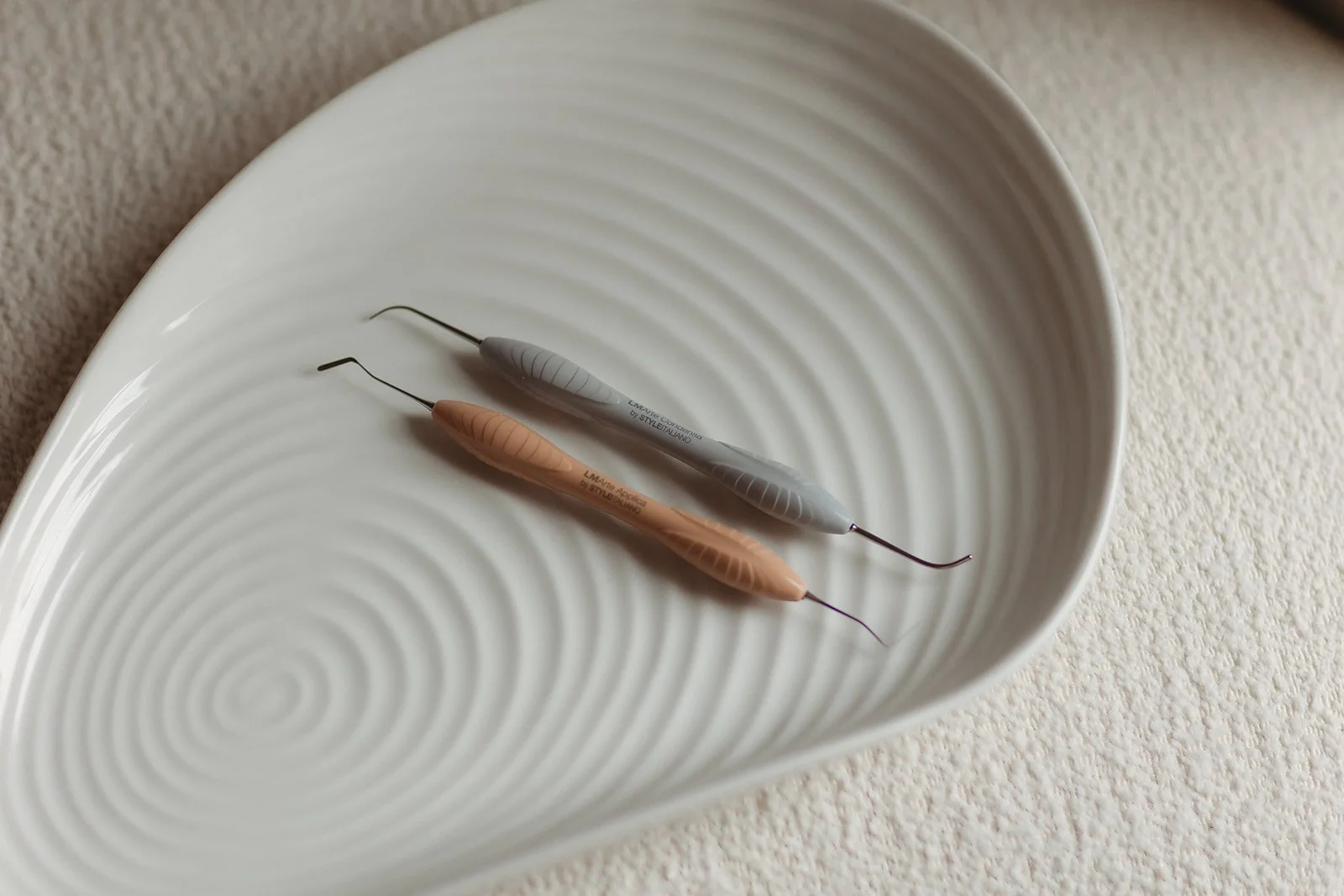 Two dental scalers from Chapter One Dental Clinic, one peach and one gray, resting inside a white, textured ceramic dish on a beige surface.