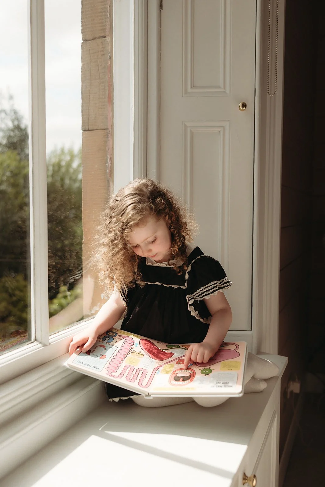 A young girl with curly hair, wearing a black dress with ruffle sleeves, sitting by a window reading a colourful children's book about dentistry. Chapter One Dental Clinic's waiting room.