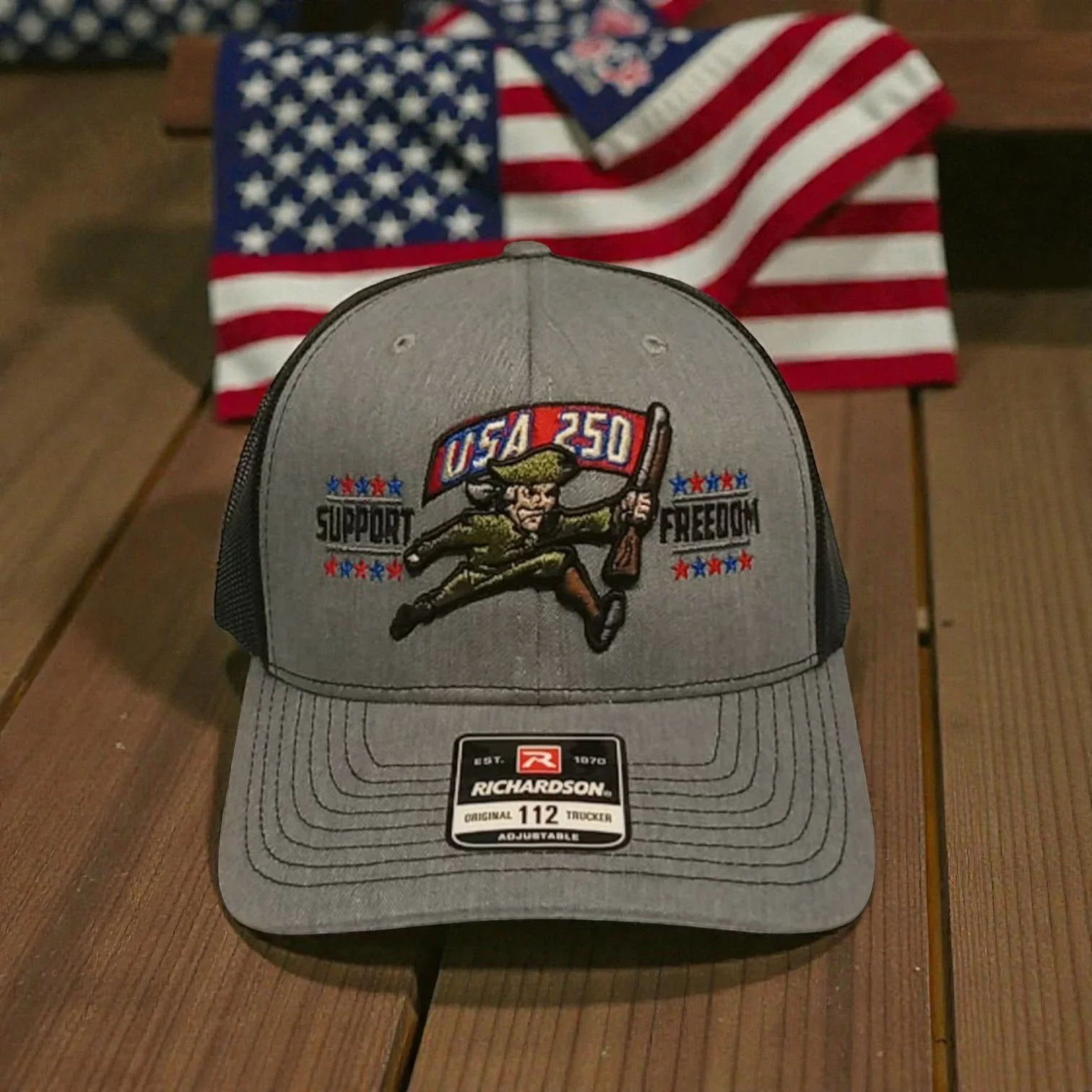 A gray Richardson trucker cap with an embroidered patch of a cartoon soldier holding a rifle and the words 'USA 250', 'Support', and 'Freedom'. An American flag is in the background.