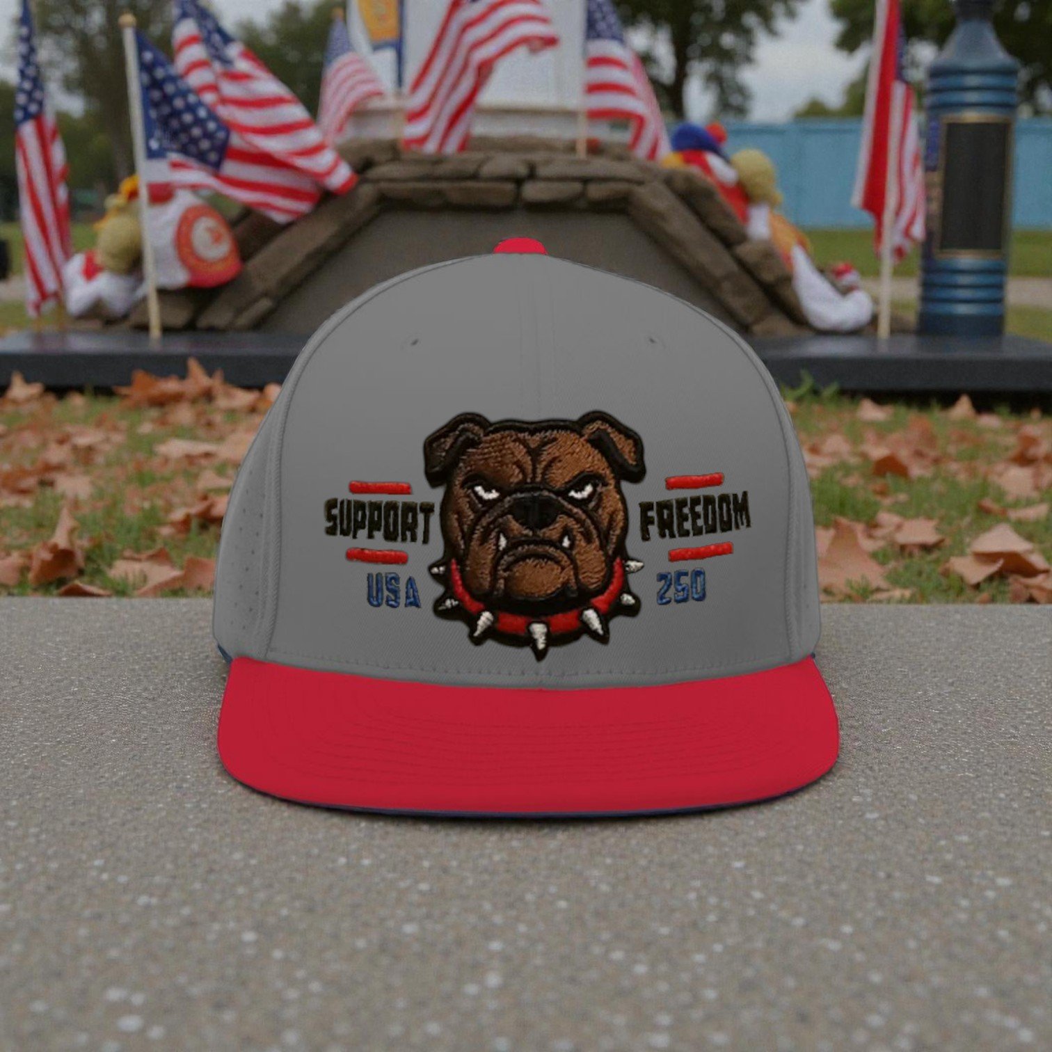 A gray and red snapback hat featuring a fierce bulldog face embroidered in the center, with the words 'Support,' 'Freedom,' 'USA,' and '250' around it, placed on a gray surface outdoors with American flags and a monument in the background.