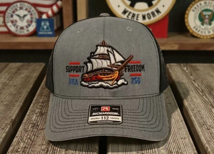 Gray trucker cap featuring embroidered image of a sailing ship on waves, with the words "Support Freedom" and "USA" on either side.