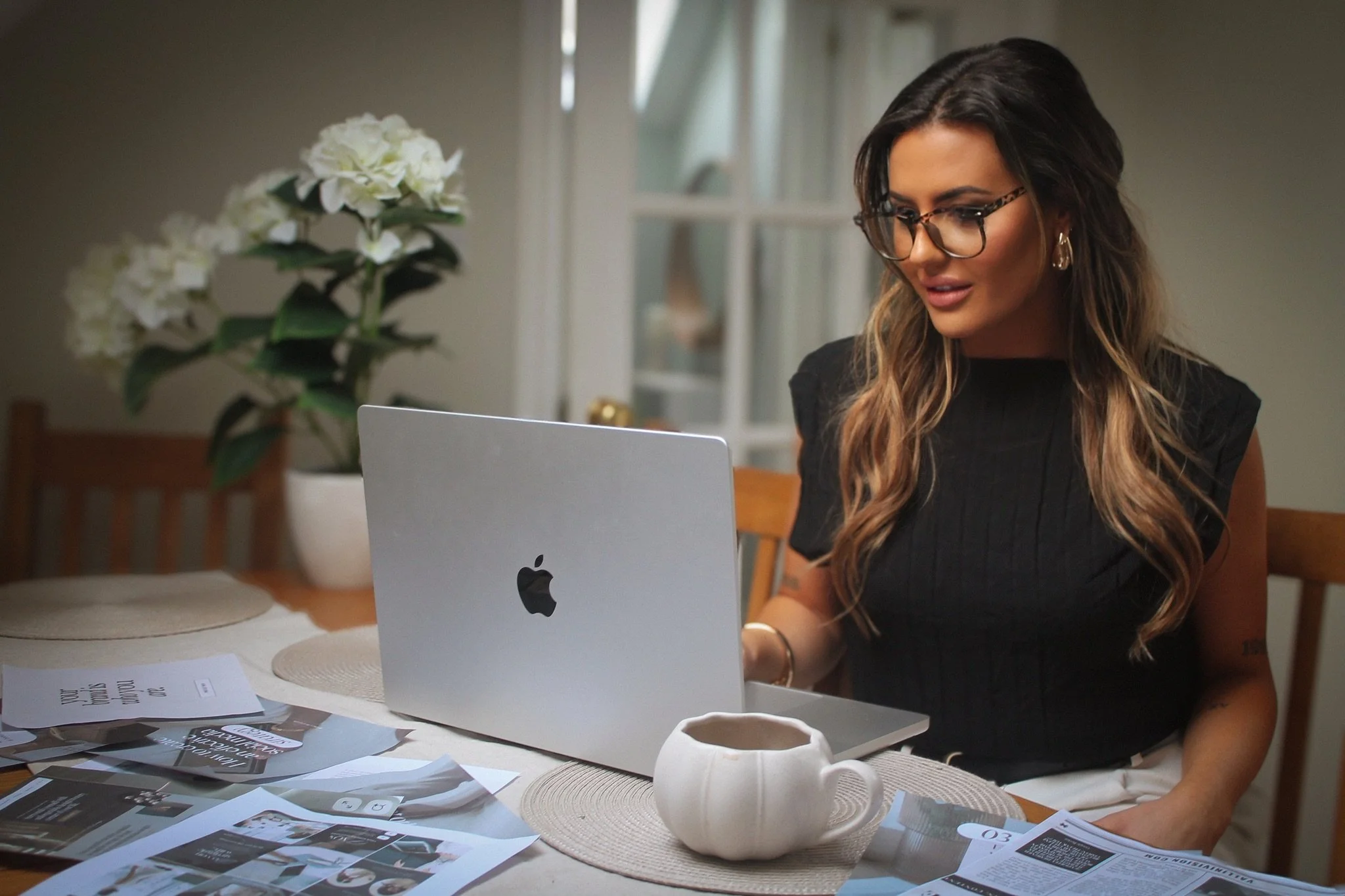 A woman with long hair, wearing glasses and a black top, sitting at a table using a silver MacBook, surrounded by papers, a white ceramic mug, a potted plant, and a window in a home interior.