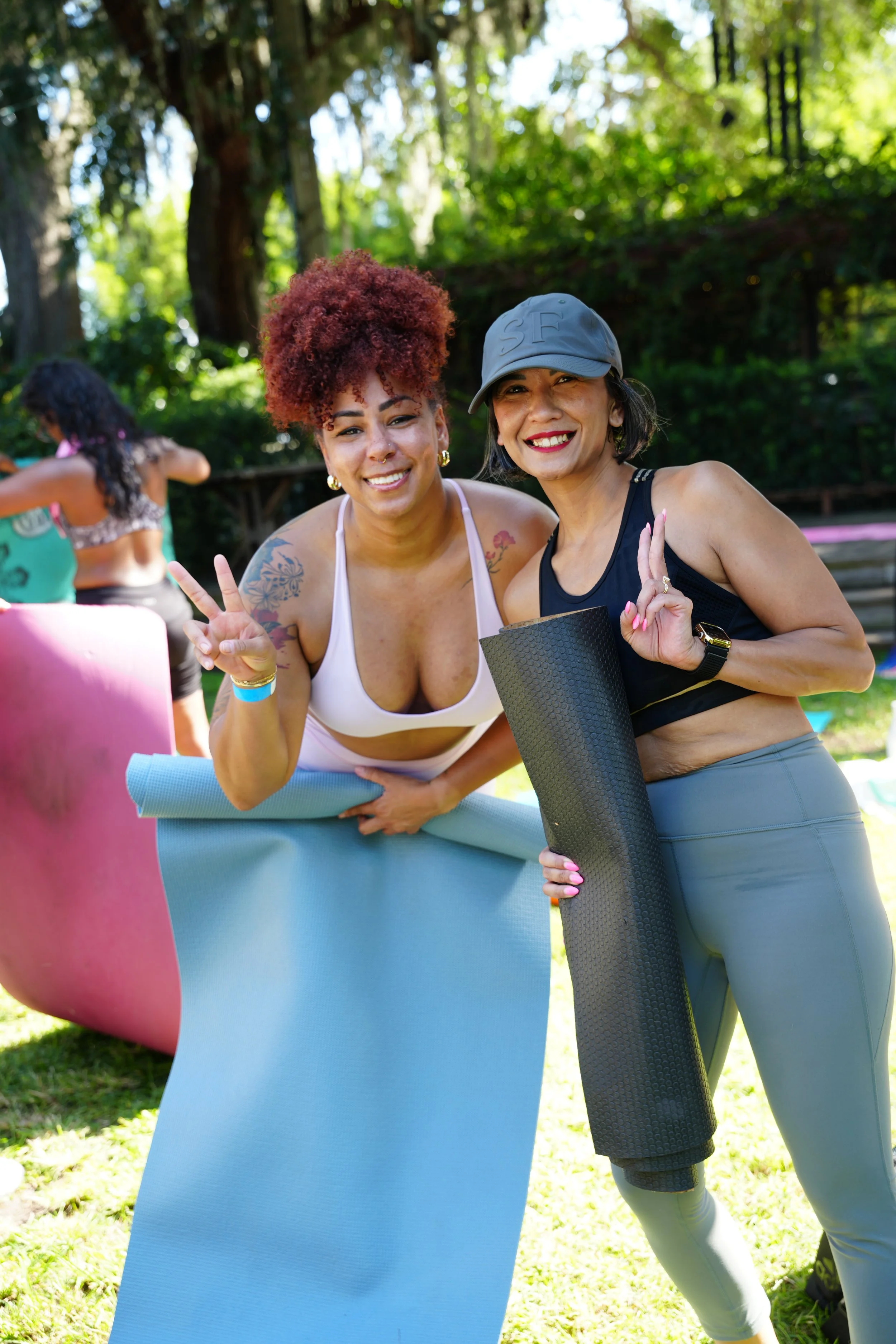 Two women smiling and posing with peace signs while holding yoga mats outdoors in a sunny, green park, with a pink yoga mat and another person in the background.