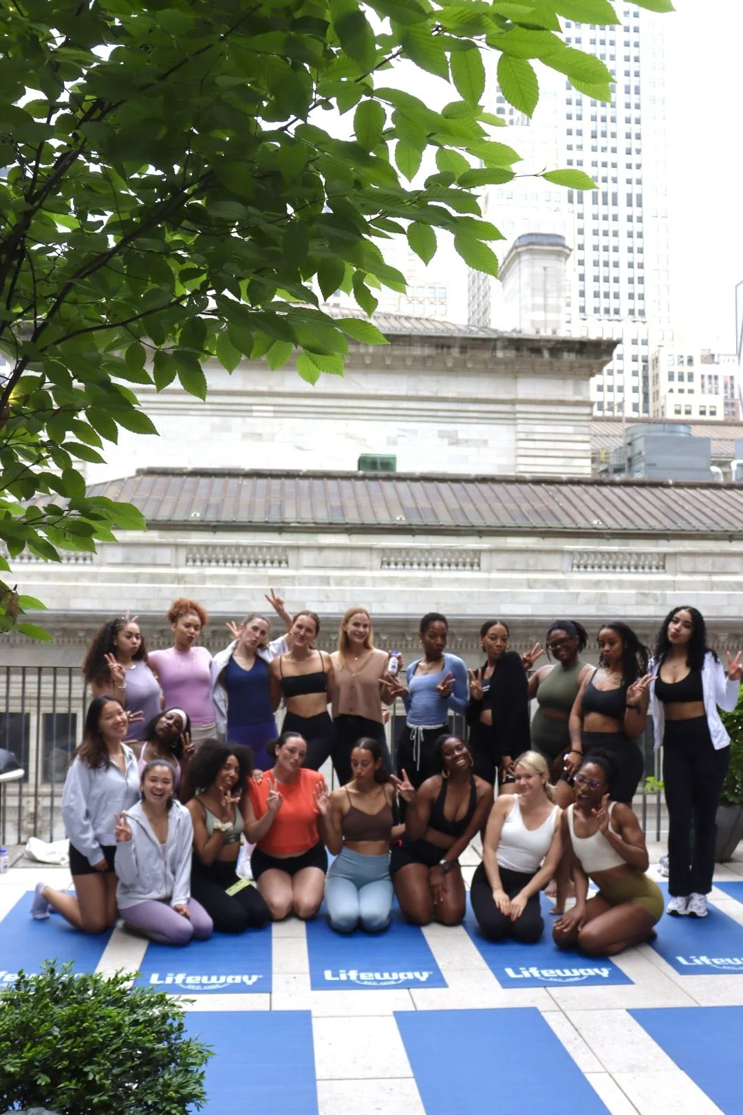 Group of diverse women practicing yoga outdoors in an urban setting, posing on blue yoga mats with skyscrapers in the background, some making peace signs and smiling.