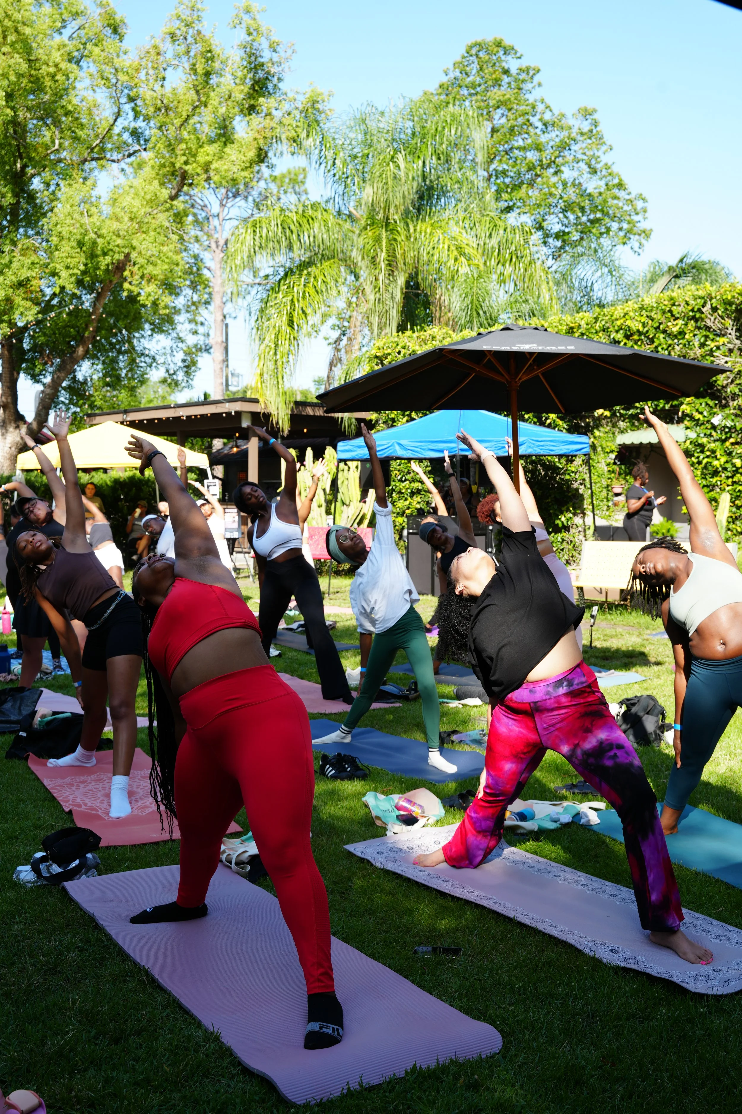 A group of people participating in an outdoor yoga or stretching class on a sunny day, with mats laid out on the grass and trees in the background.