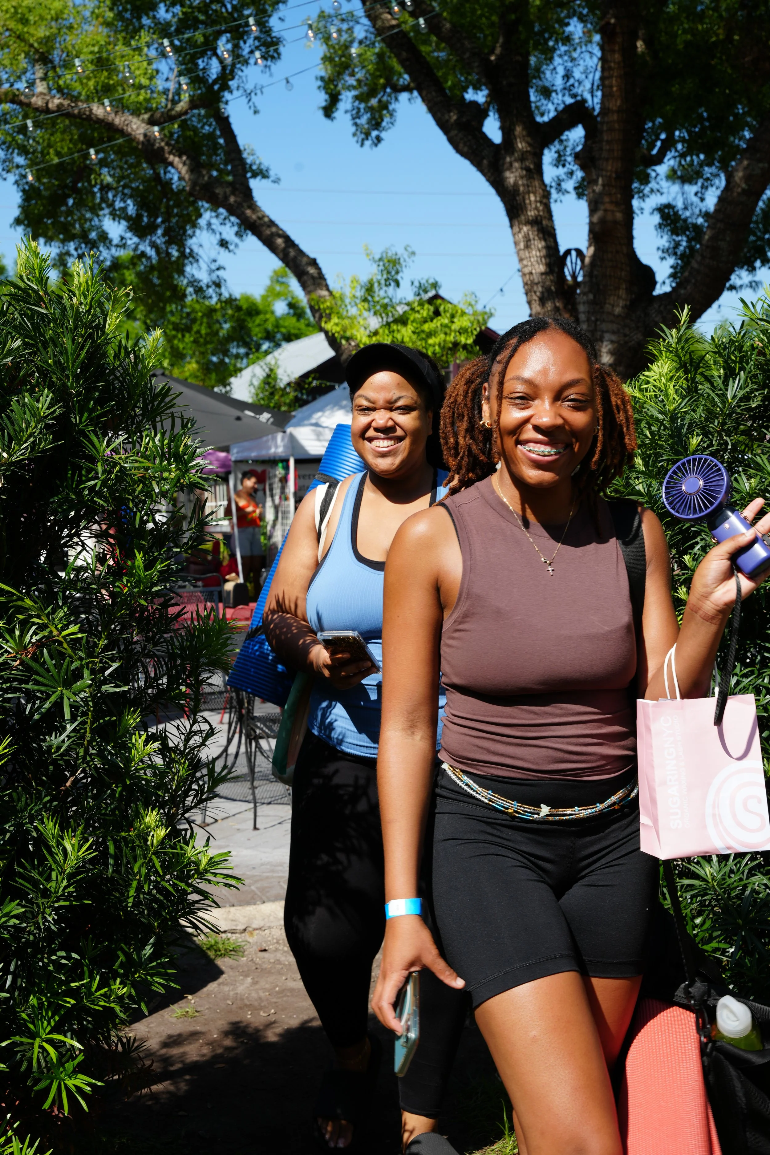 Two women smiling outdoors, walking through a garden area with tents and trees in the background, one holding a portable fan and a pink shopping bag.