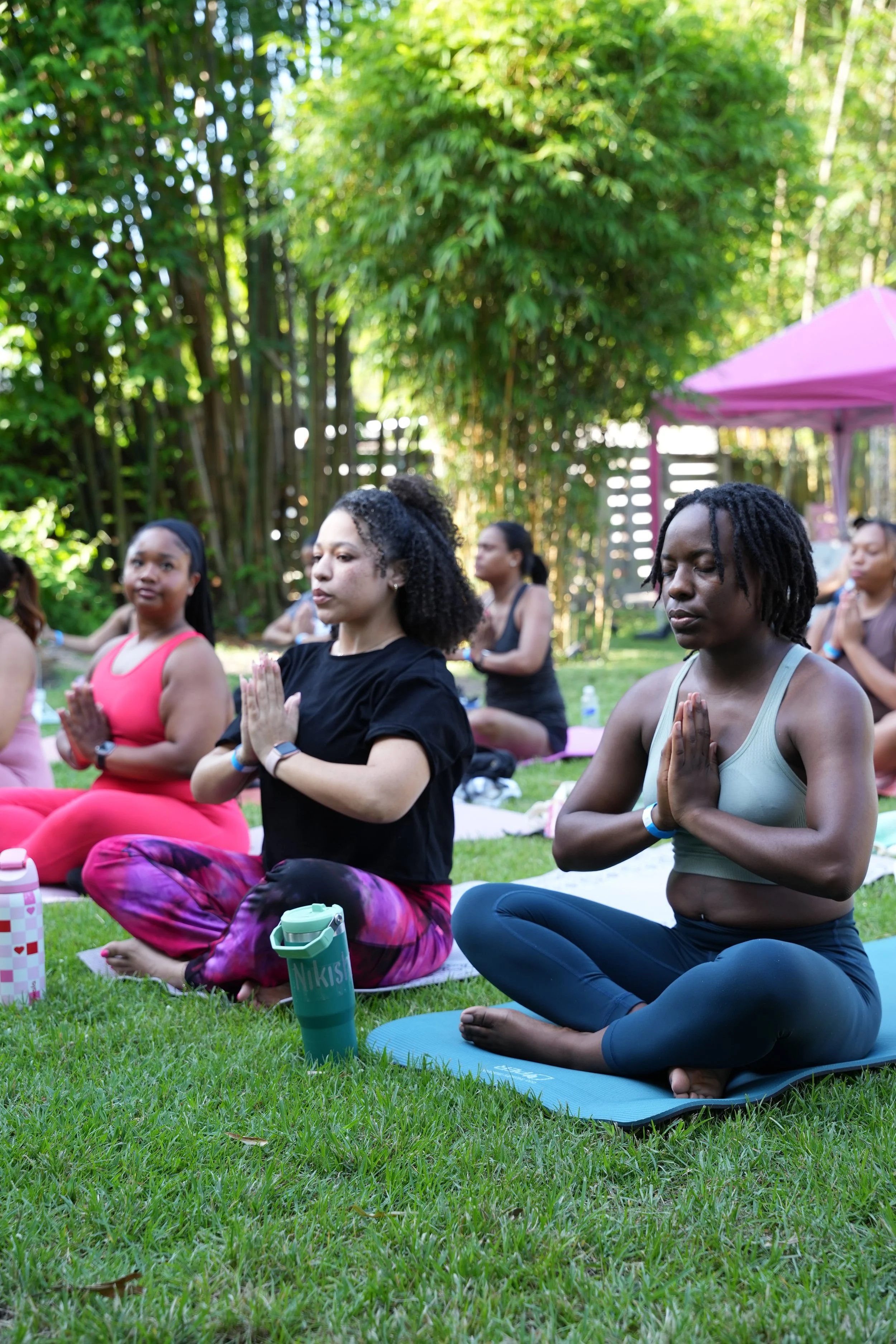 Group of women practicing yoga outdoors on a grassy area surrounded by trees, sitting in a cross-legged meditation pose with hands in prayer position.