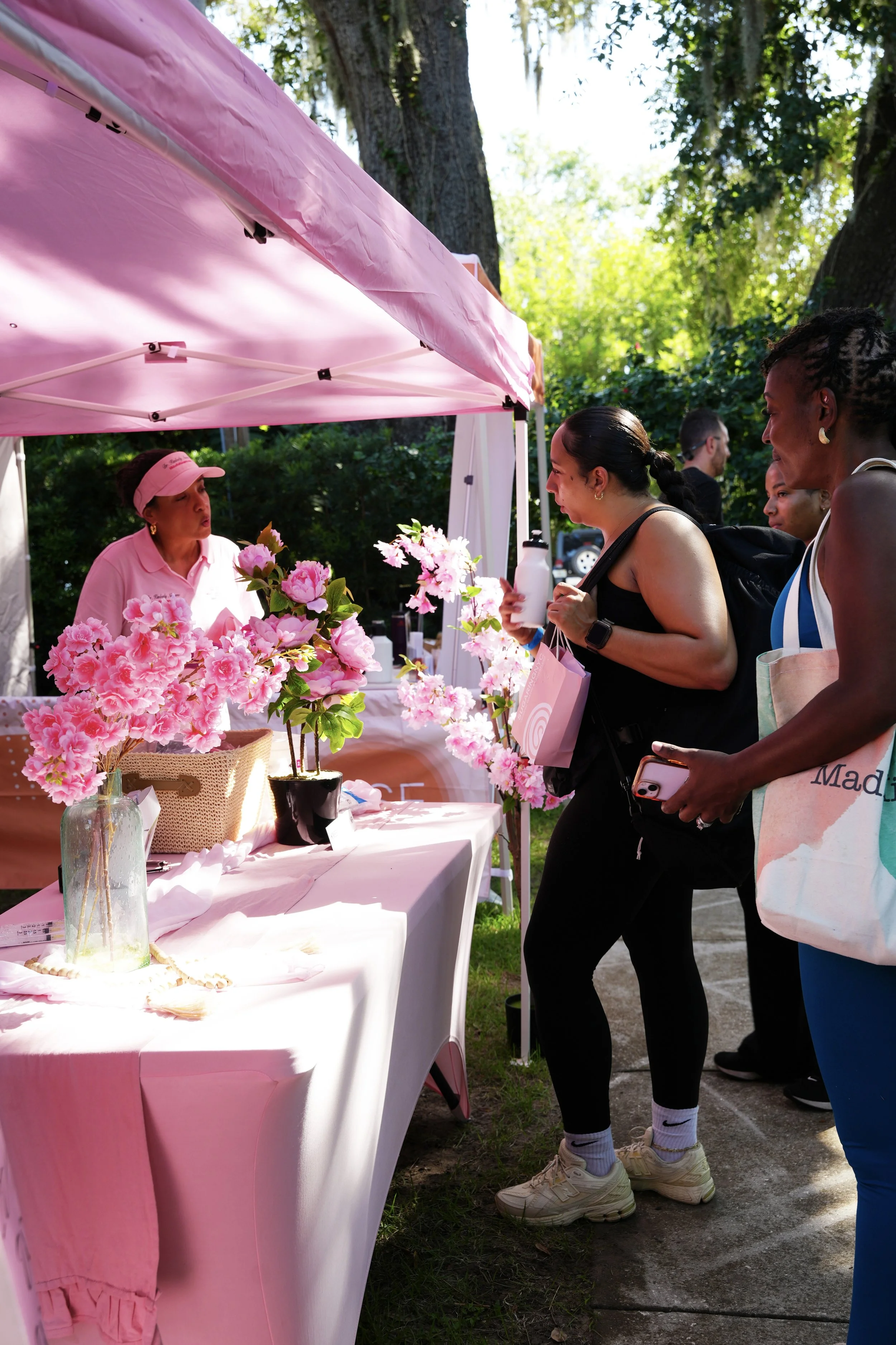 Women shopping at a pink floral booth at an outdoor market or fair.