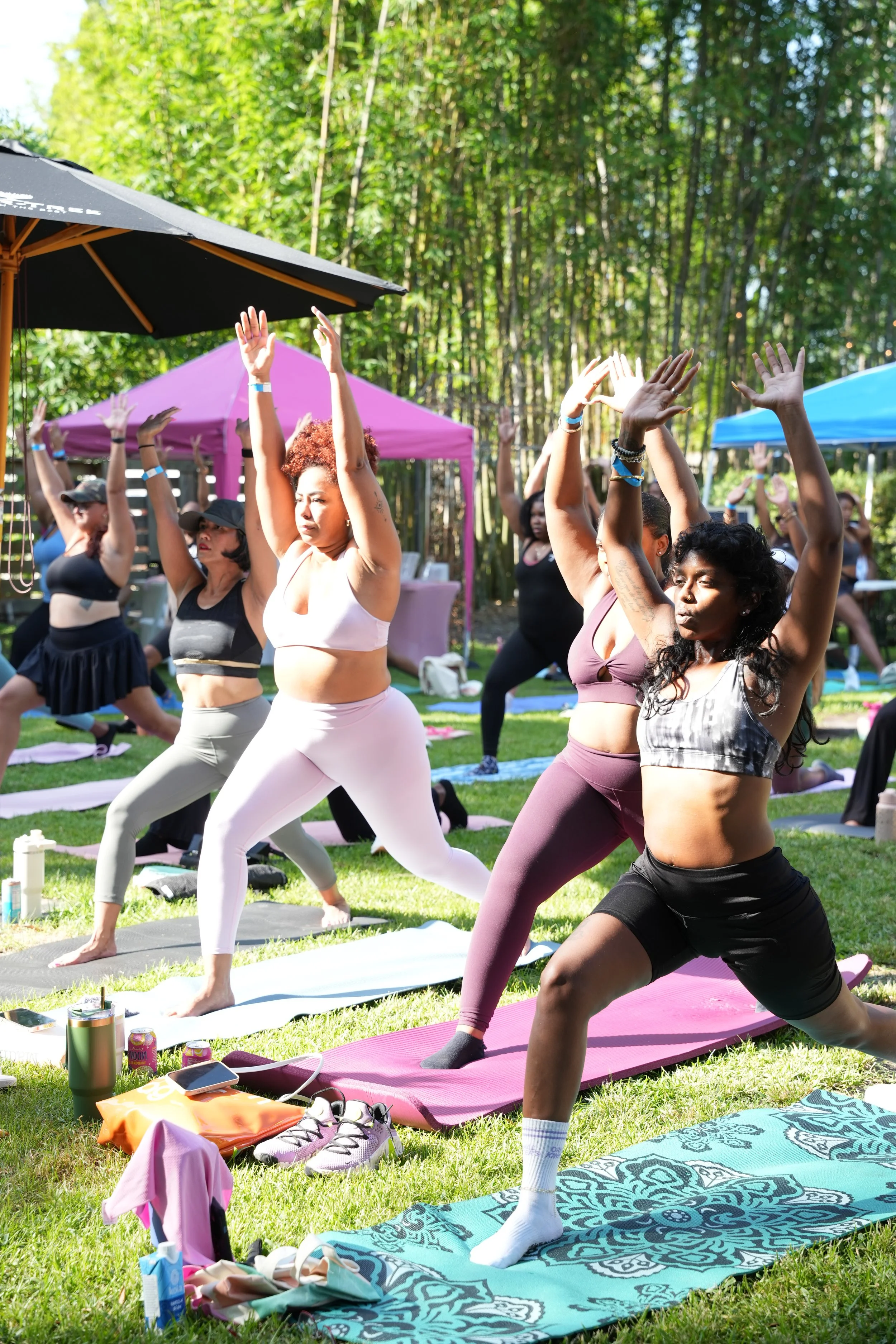 A group of women practicing yoga outdoors in a grassy area with trees in the background, under colorful tents, during daytime.