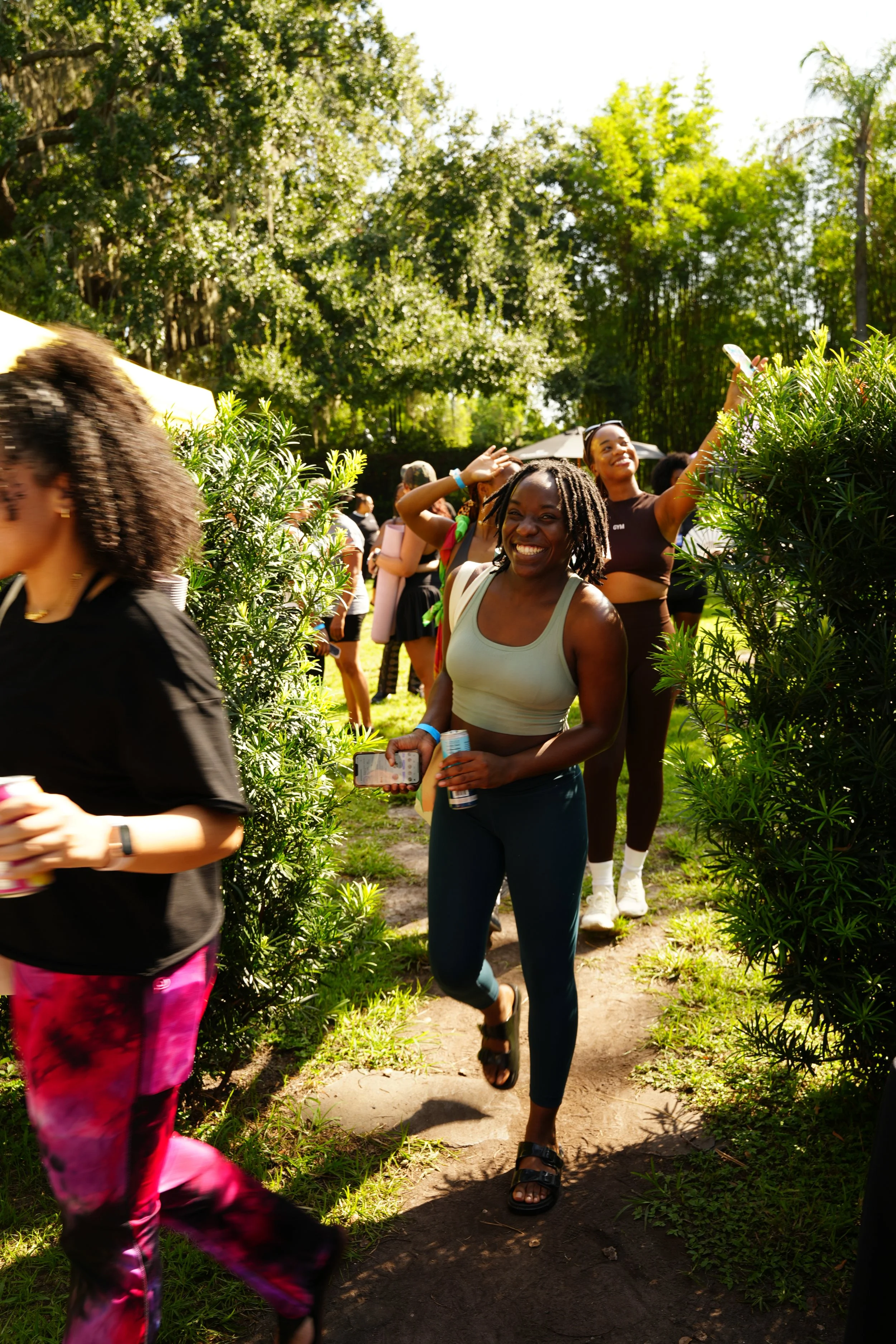 Smiling woman in athletic wear walking through a green outdoor park during daytime, surrounded by other people enjoying the outdoors.