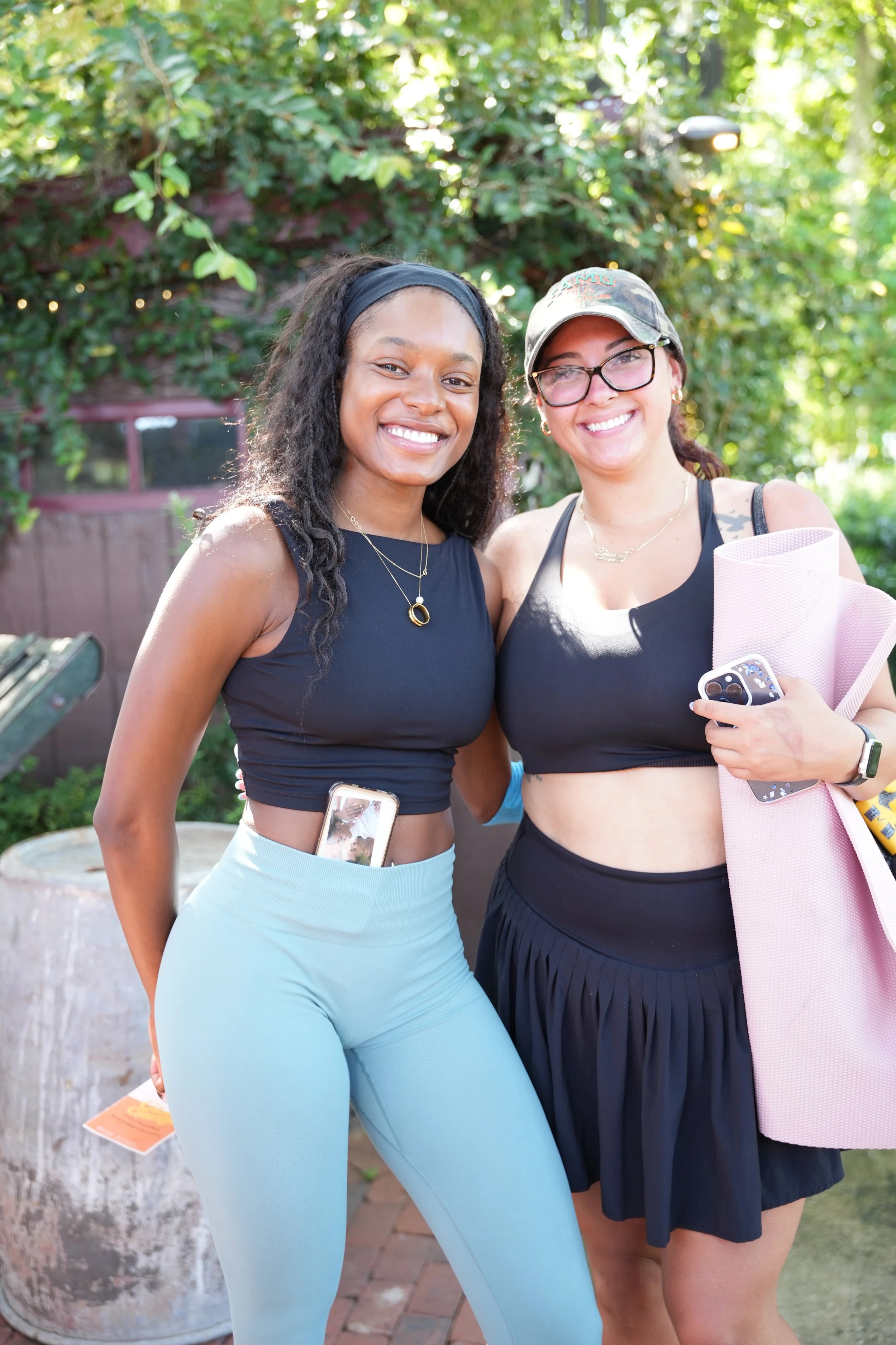 Two women smiling outdoors, one wearing light blue leggings and a black tank top, the other in a black skirt and sports bra, holding pink rolled-up mat and cell phone, standing on brick patio with greenery background.