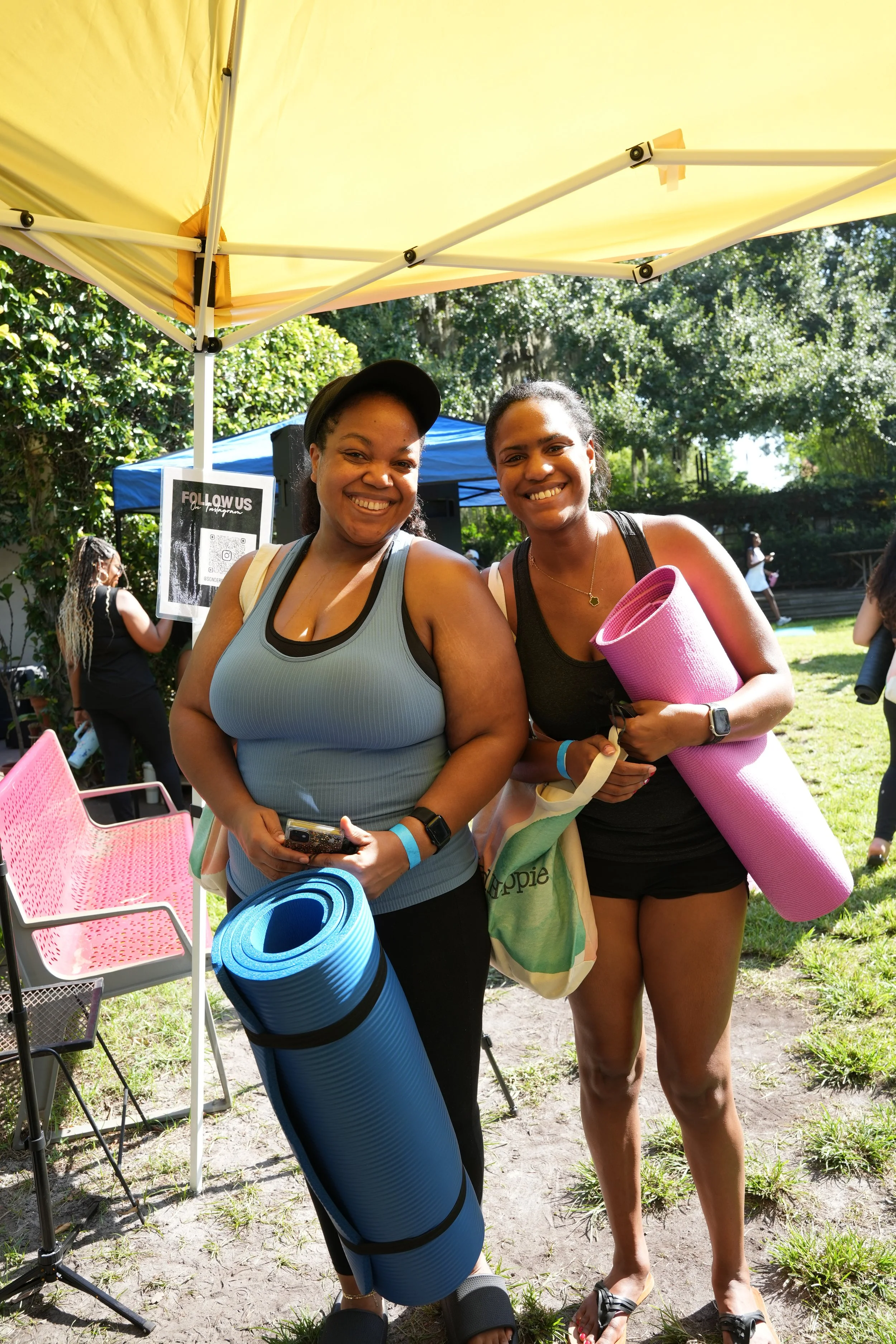 Two women smiling at an outdoor yoga event, each holding colorful yoga mats, standing under a yellow canopy with other people and trees in the background.