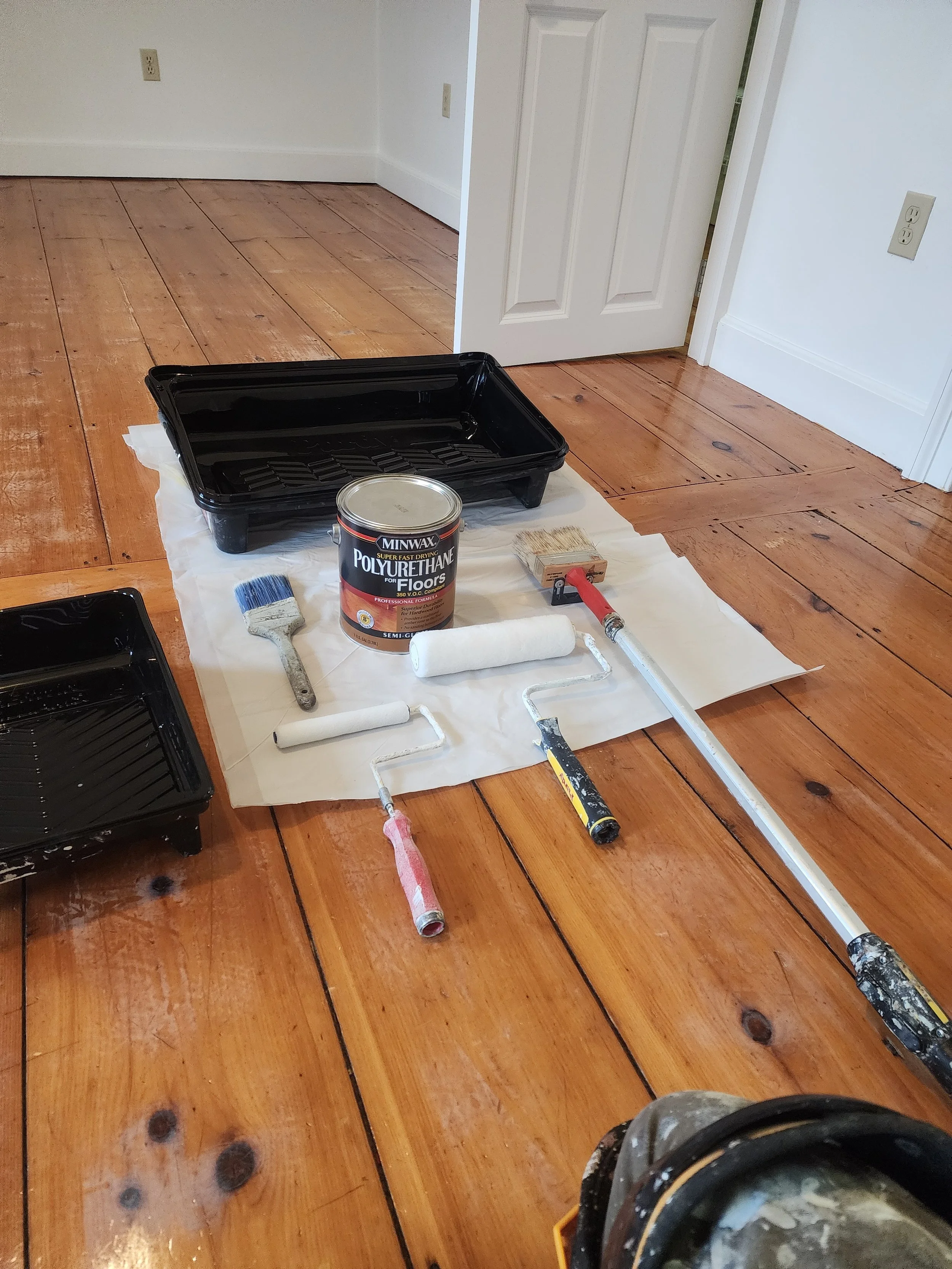 Room with wooden floor, open door, painting supplies including black paint tray, roller brushes, paint can, and small paint roller, placed on a white sheet.