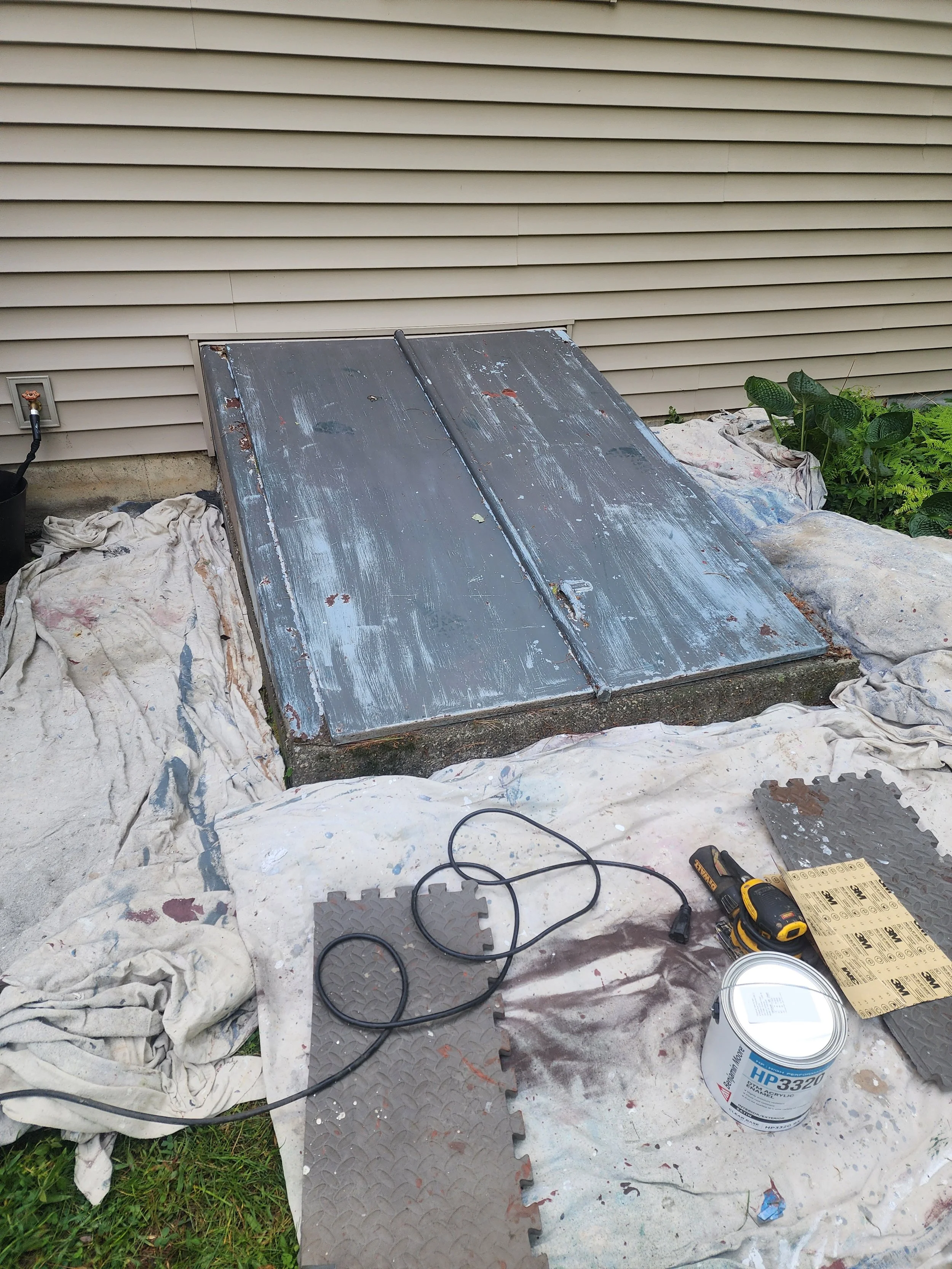 A small outdoor workspace with a weathered metal hatch door on a concrete slab, surrounded by painter's drop cloths, work tools, and paint supplies, with beige house siding and some greenery in the background.