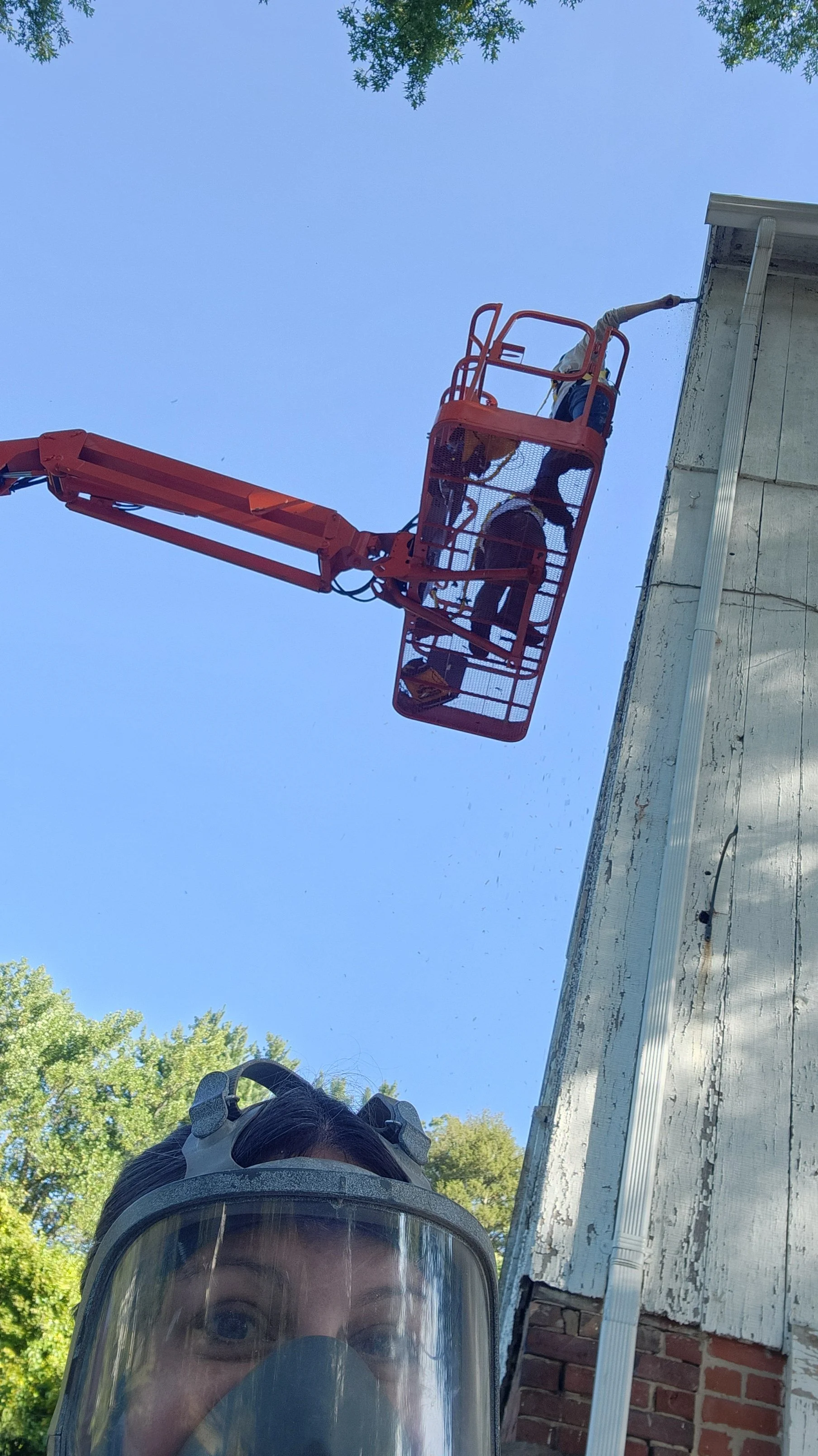 Photograph of a person in a safety helmet and face mask taking a selfie while workers are elevated on a cherry picker working on the exterior of a building under a clear blue sky.