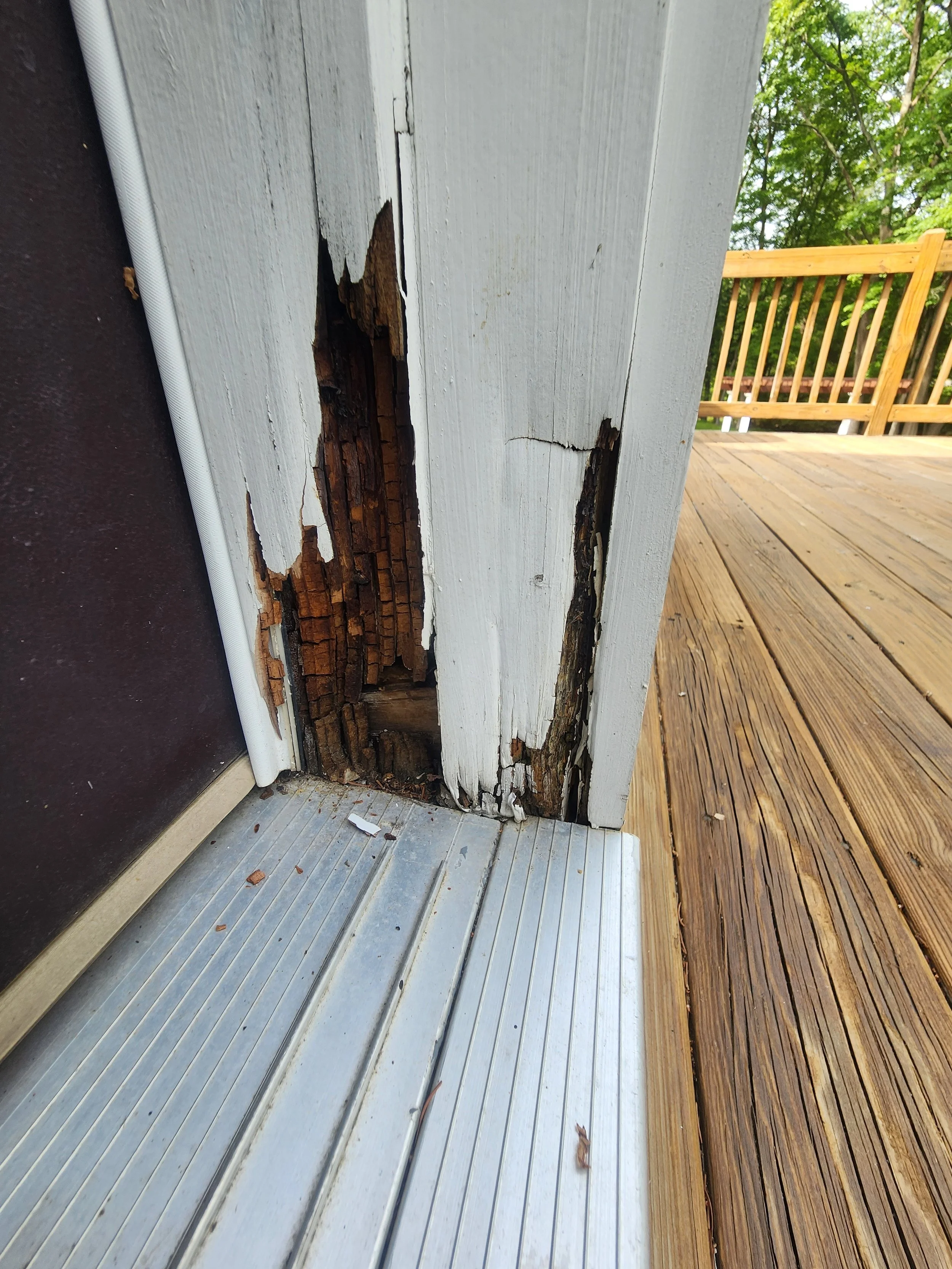 Close-up of a wooden door frame with significant rot and damage, showing peeling white paint and exposed, decayed wood.