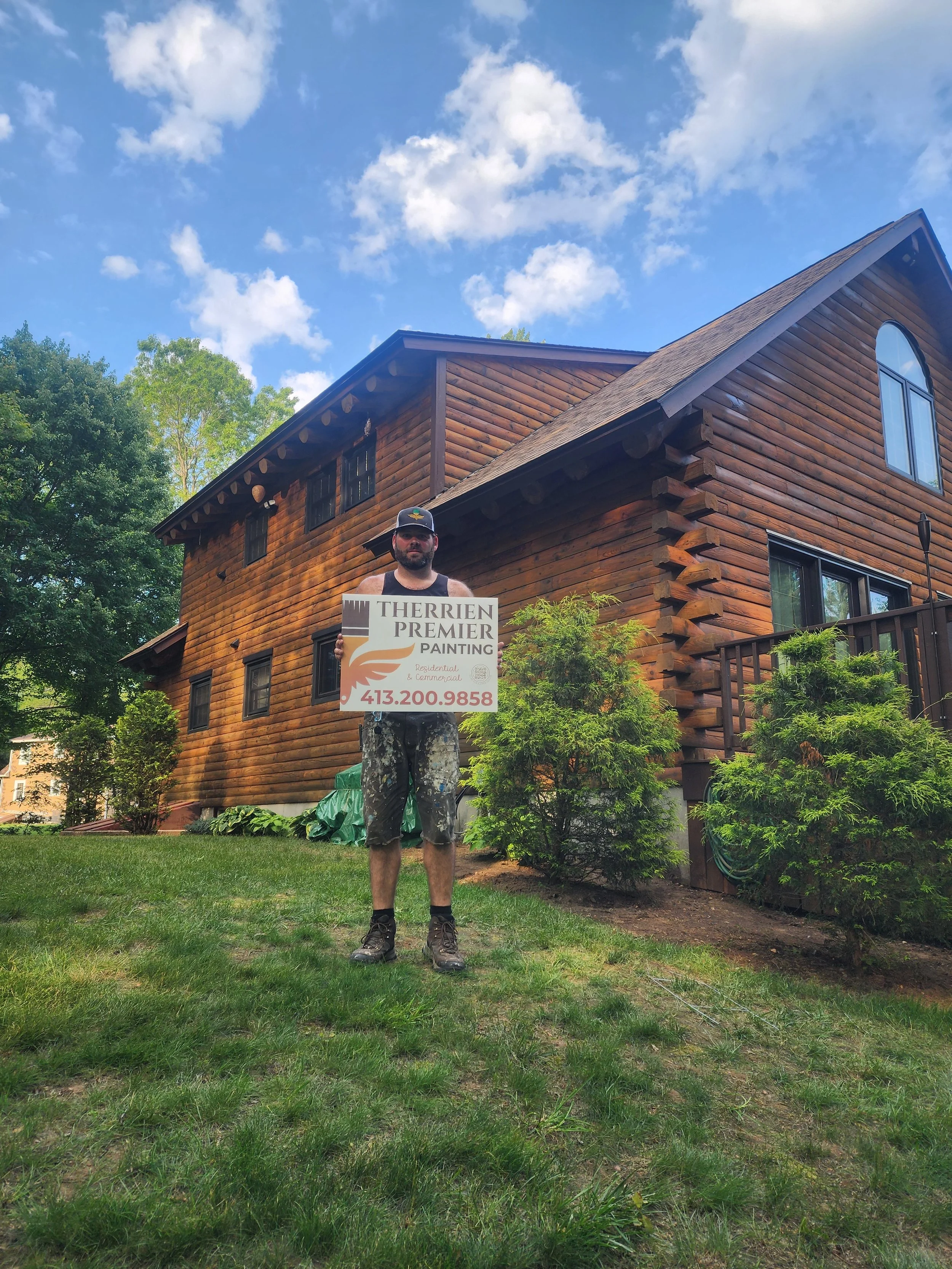 A man holding a sign for Therrien Premier Painting outside a wooden house on a sunny day.