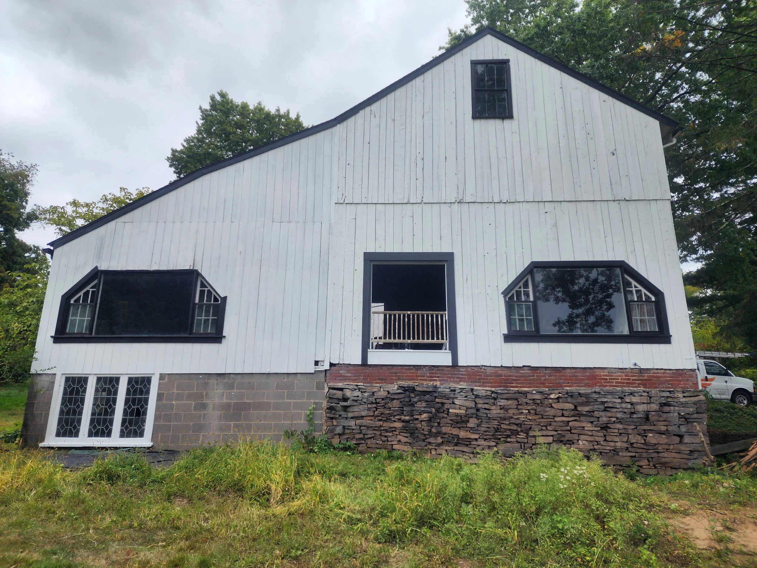 A white barn with black window frames, a small balcony, and a stone foundation, surrounded by greenery, under a cloudy sky.