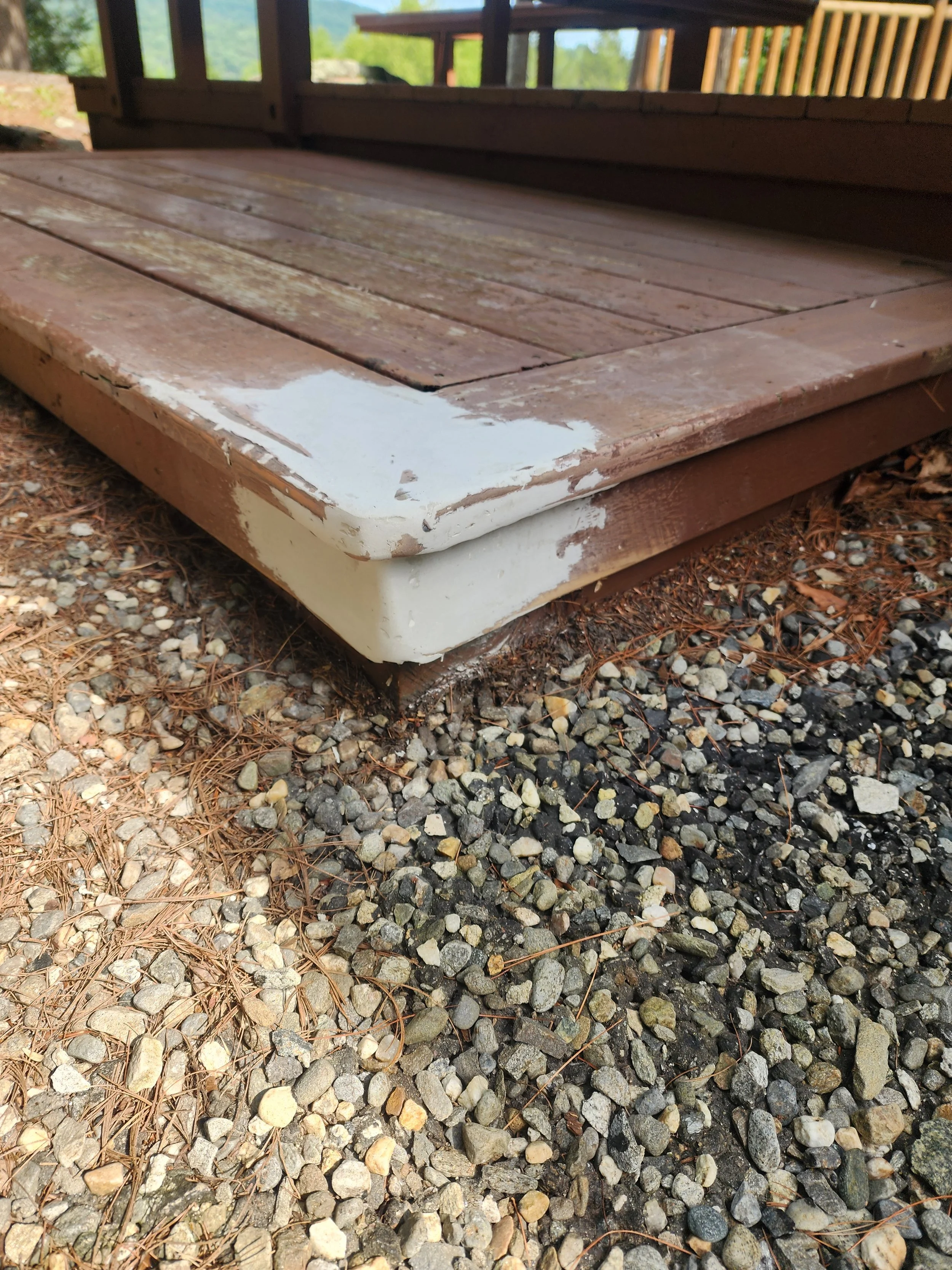 Close-up of a weathered wooden deck with peeling white paint on its edges, resting on gravel and pine needles outdoors.