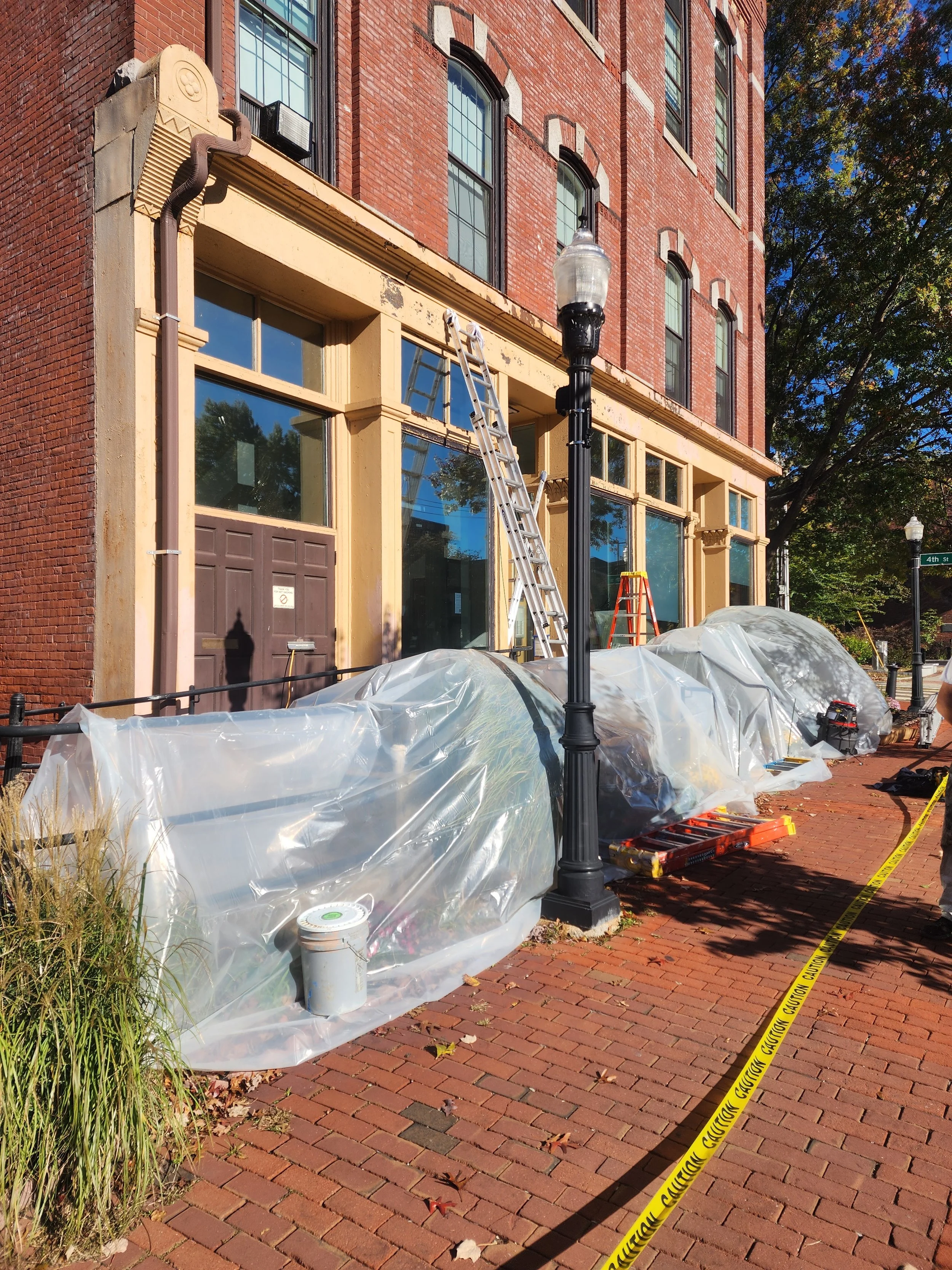 Construction work on a brick building sidewalk, with plastic covers over plants, ladders, tools, and caution tape.