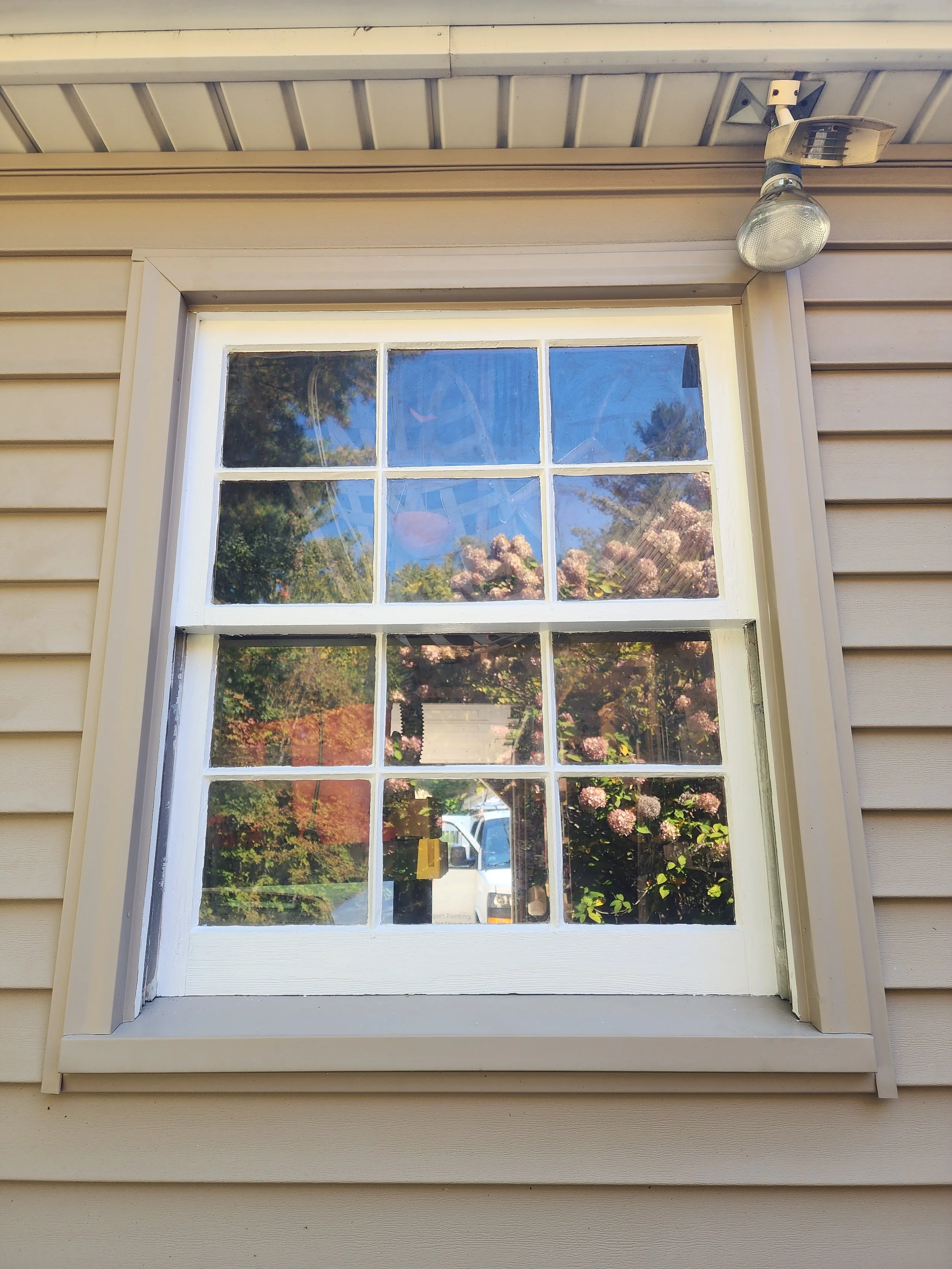 A window with six panes on a beige house wall, reflecting trees, pink flowers, blue sky, and a white vehicle outside. A security light is mounted above the window.