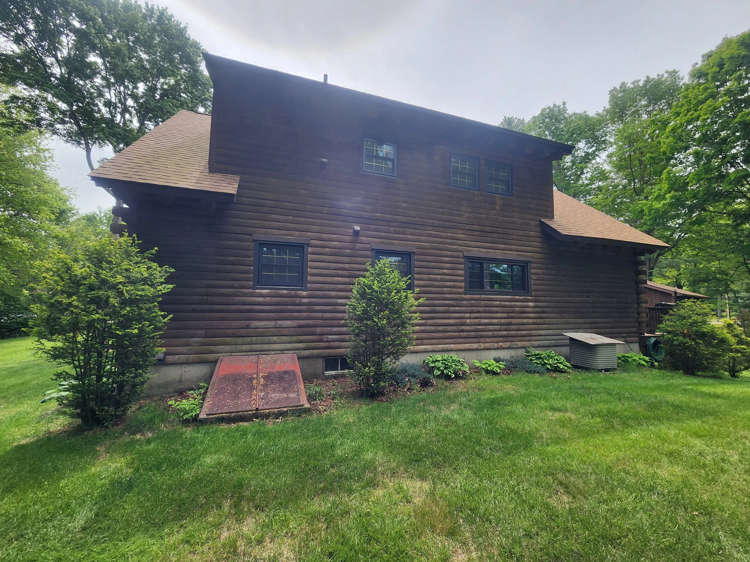 A rustic wooden house with a sloped roof, surrounded by green trees and grass.