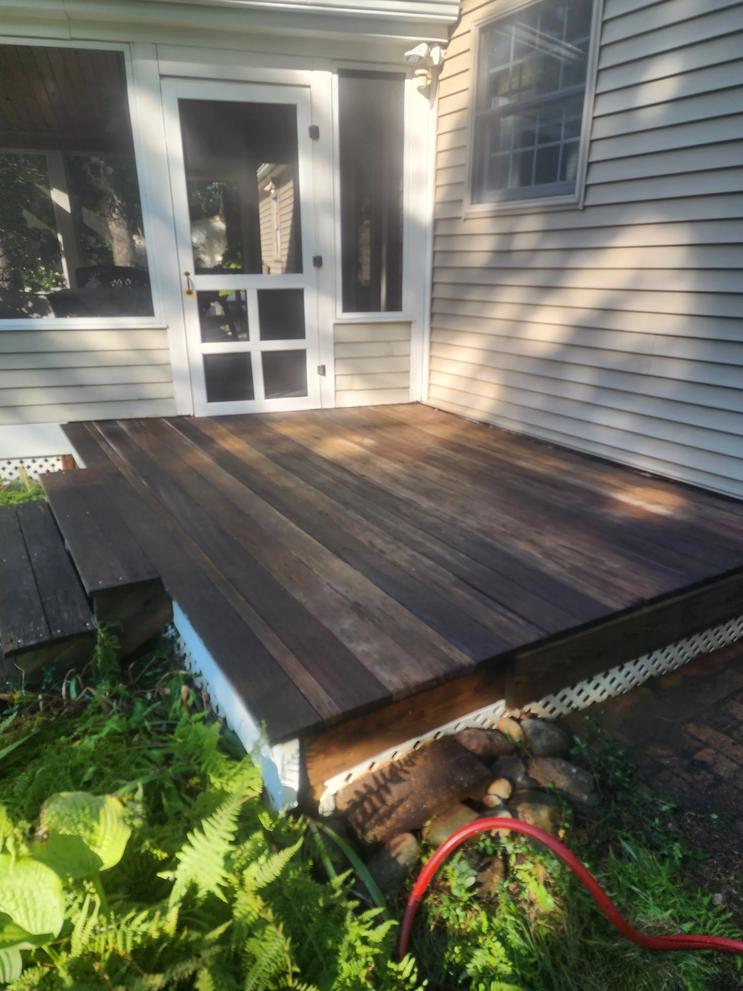 A newly built wooden deck attached to a house with white siding, near a screened porch door, with some greenery and rocks at the base.