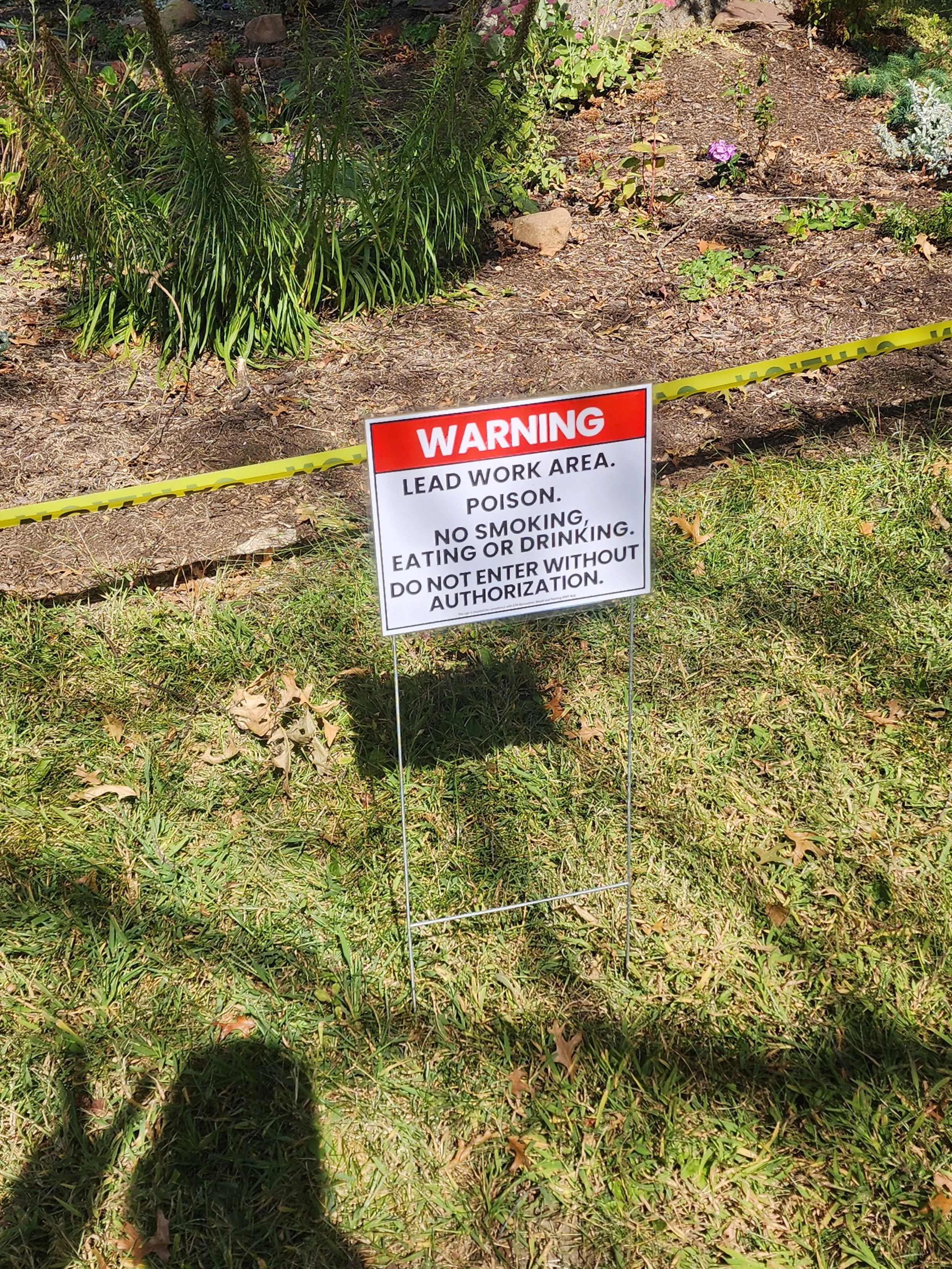 Warning sign on a grassy area indicating a lead work zone with poison, no smoking, eating, or drinking, and no entry without permission.