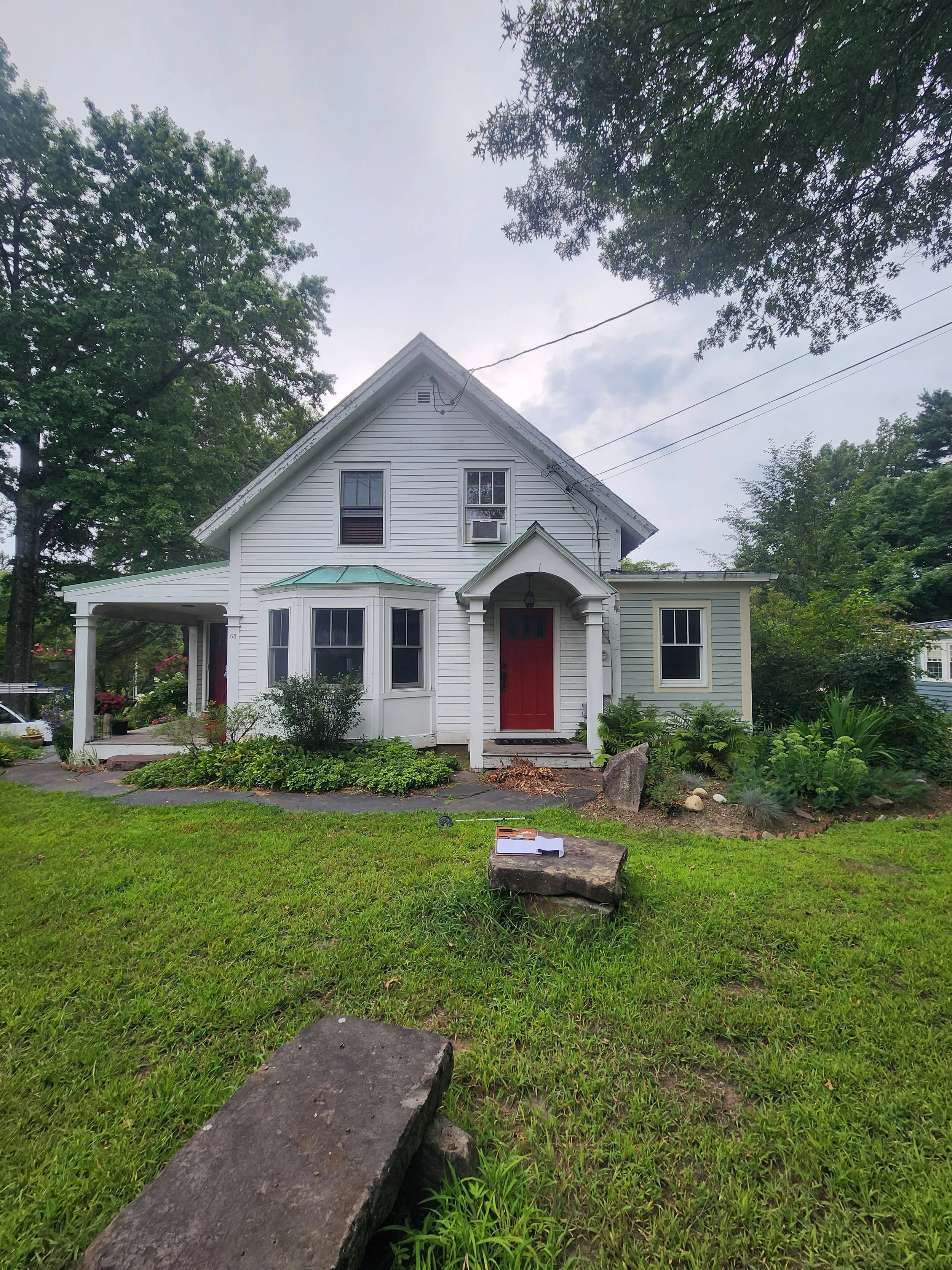 A two-story white house with a red front door and a porch, surrounded by a well-maintained lawn and trees, with cloudy sky overhead.