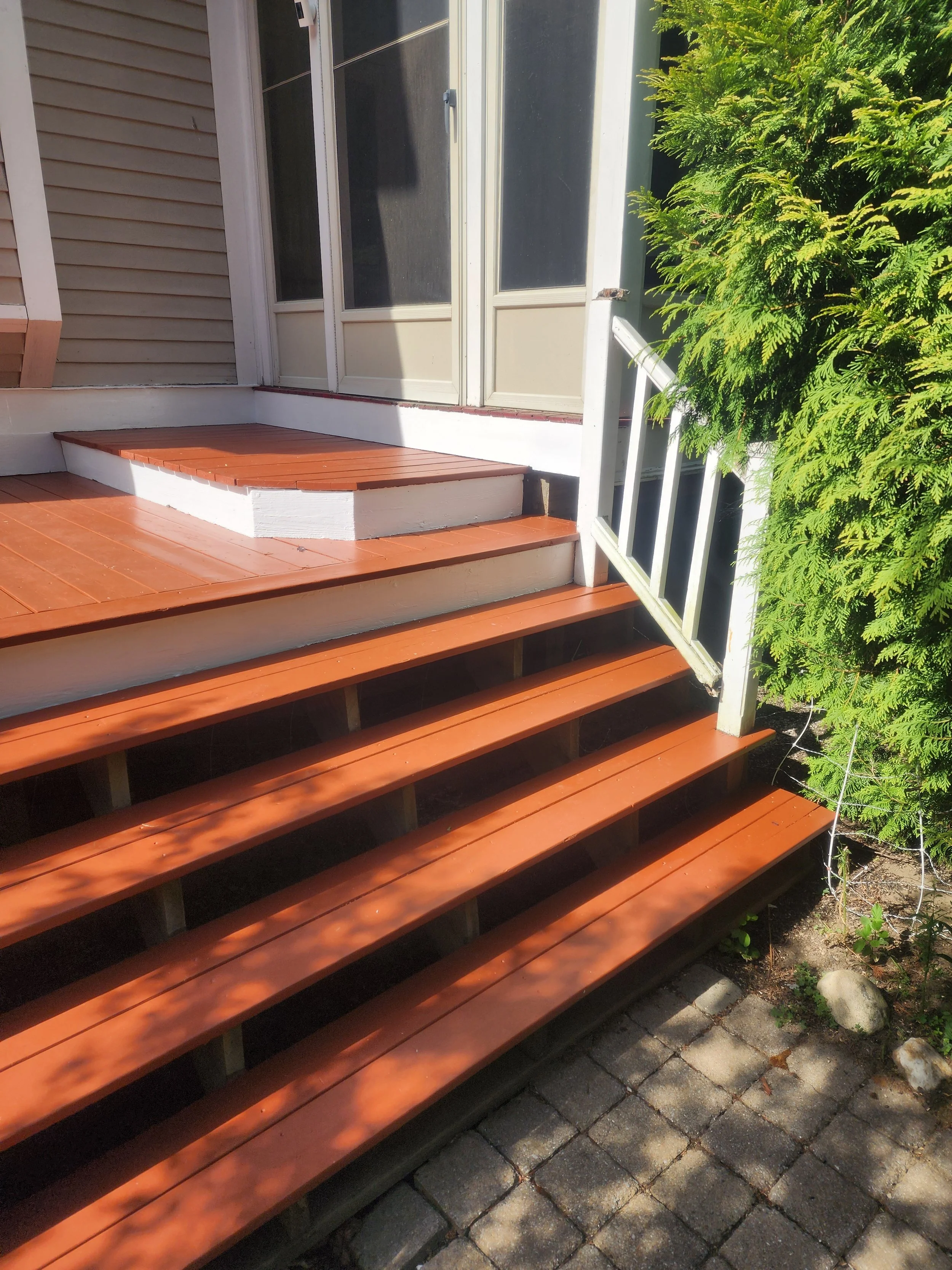Image of a wooden porch staircase with newly stained steps leading up to a screened door on a house, with a green bush on the right side and a stone pathway below.