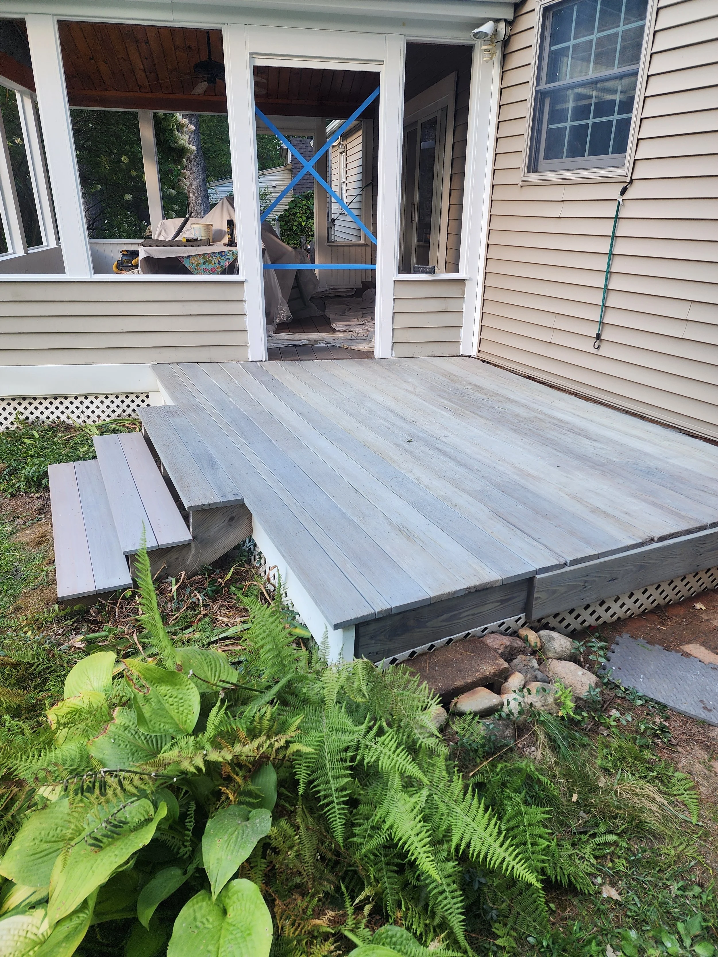 Backyard view of a screened porch with a wooden deck, partially under construction, with blue tape crossing the door frame, surrounded by green plants and garden rocks.