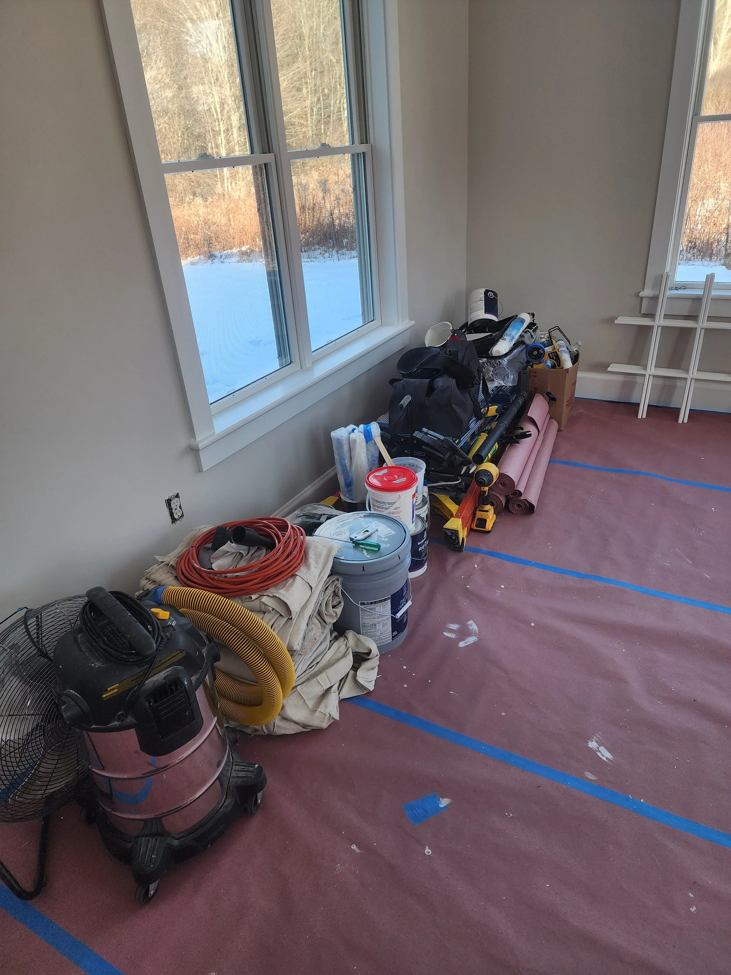 A room under renovation with construction tools and materials along the wall beneath large windows, with a snowy outdoor landscape visible outside.