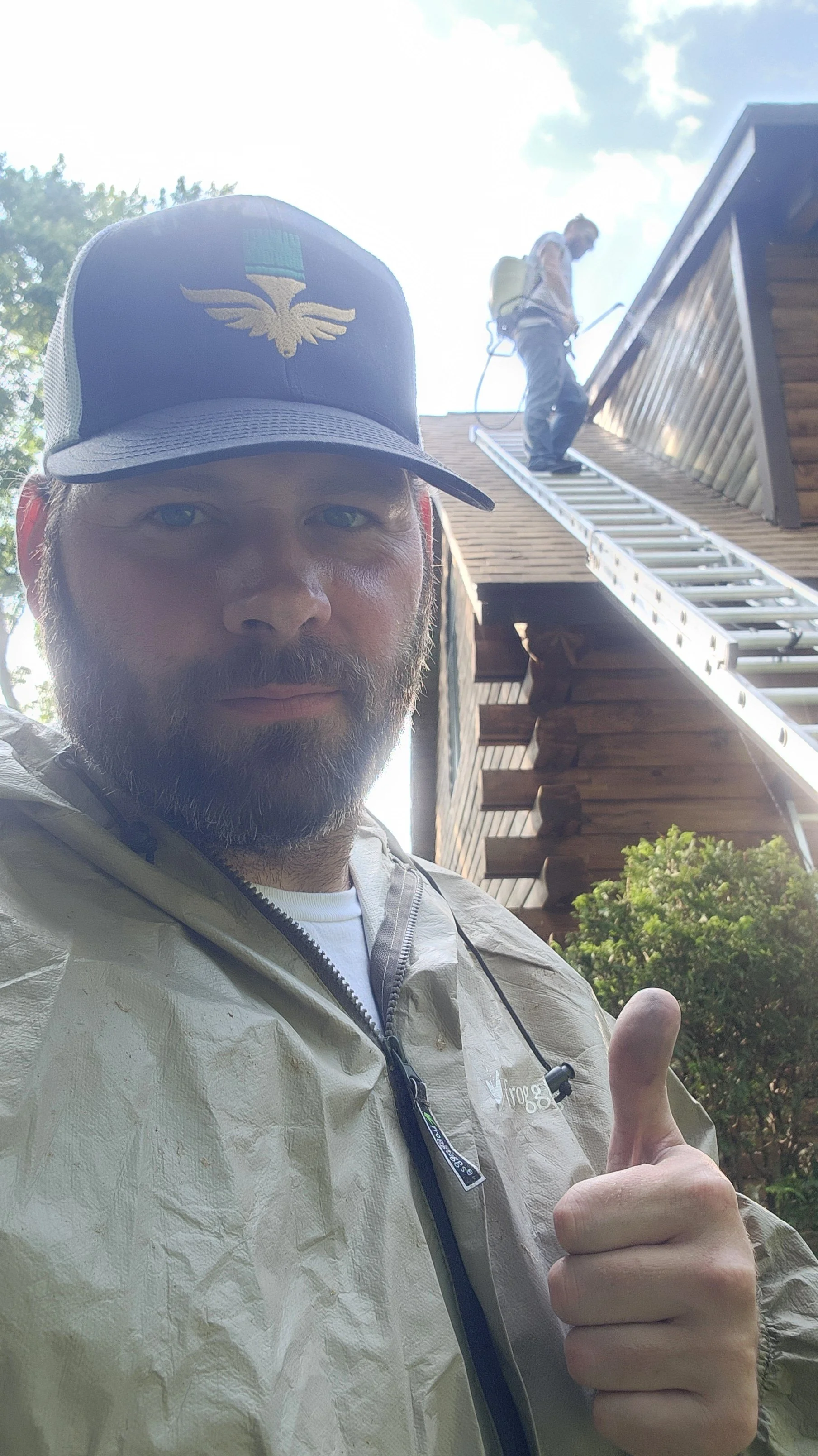 Man with a beard and a trucker hat giving a thumbs-up in front of a house with a person on the roof carrying out maintenance.