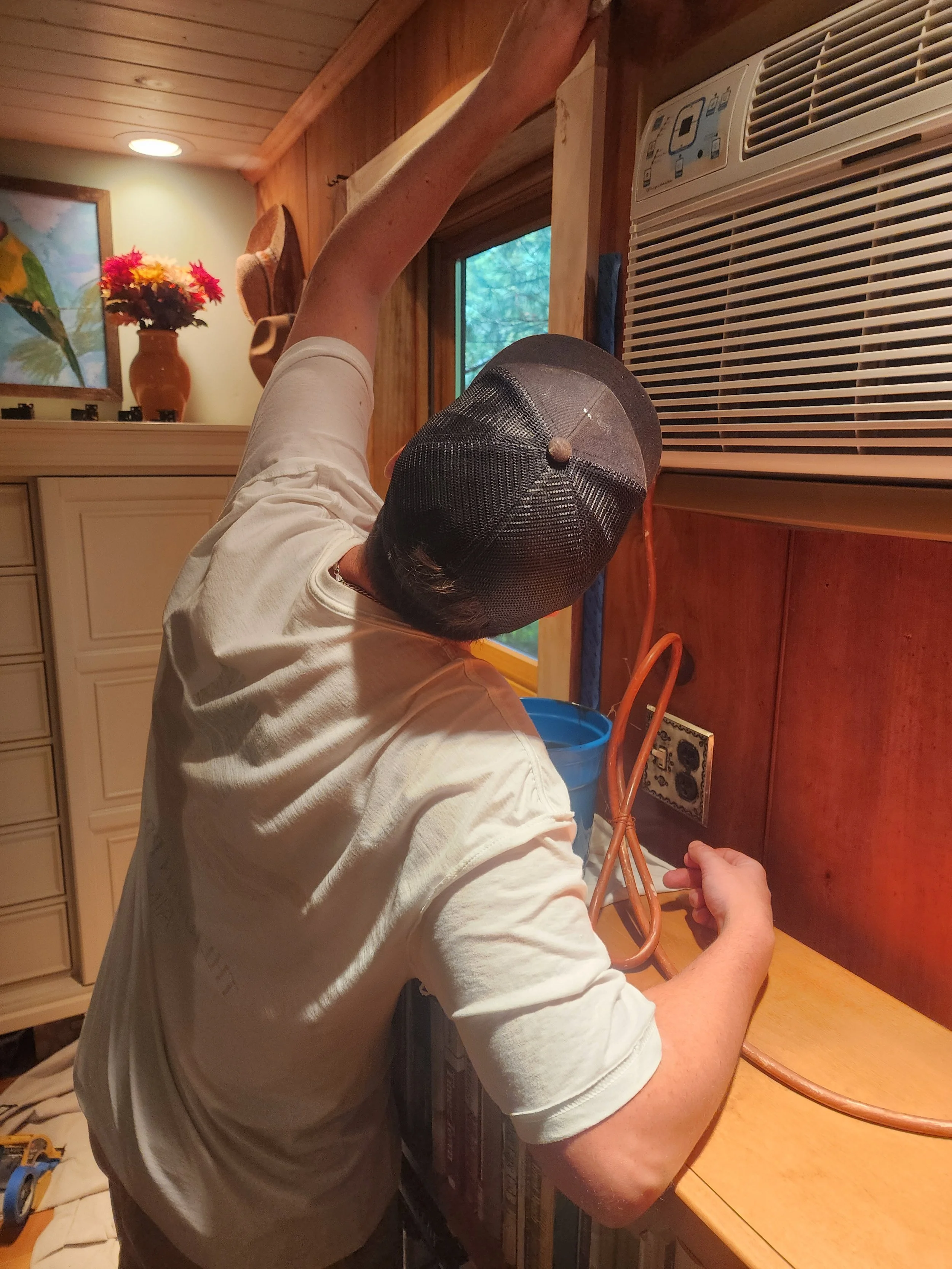 A person working on wiring next to an air conditioning unit in a wooden-paneled room.